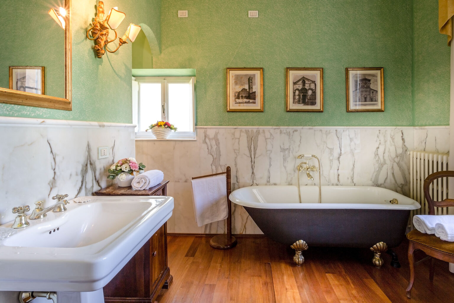 Elegant vintage bathroom featuring a clawfoot bathtub, a decorative sink with antique faucet fixtures, and a wooden cabinet. The walls are adorned with green wallpaper and marble accents, complemented by framed artwork and a window allowing natural light. A wooden chair and neatly rolled towels add to the inviting atmosphere. | Only on SopranoVillas