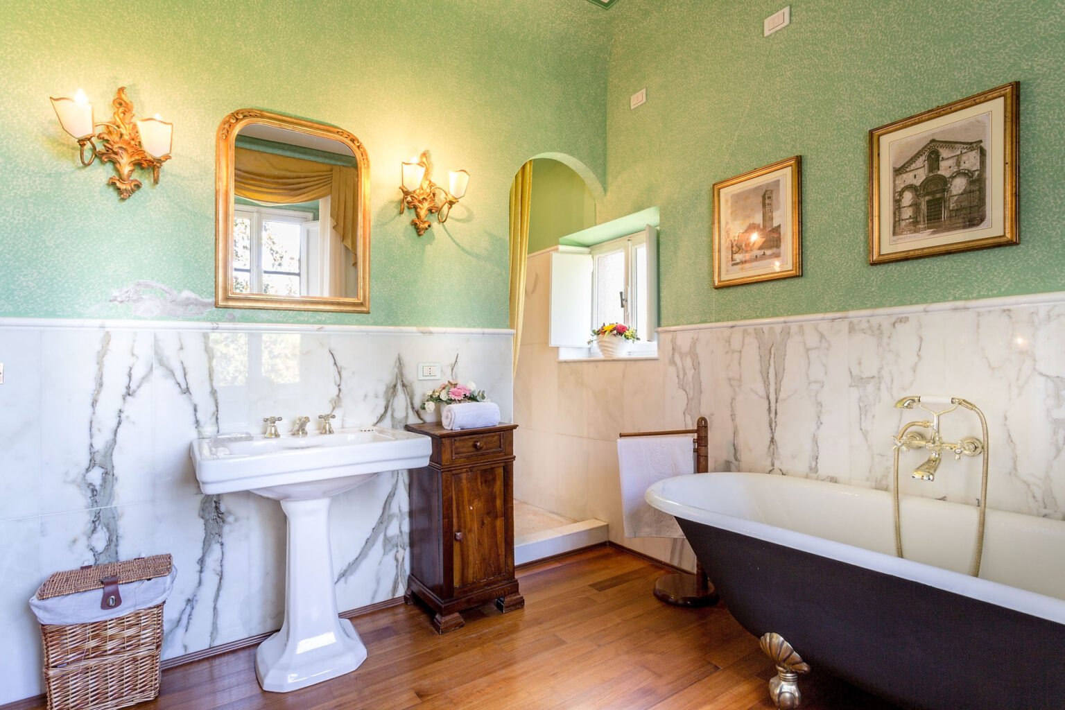 Elegant vintage bathroom featuring a green textured wall, marble accents, a classic pedestal sink, and a black freestanding bathtub. The space is adorned with decorative wall sconces, framed artwork, and natural light from a window, creating a serene and stylish atmosphere. | Only on SopranoVillas
