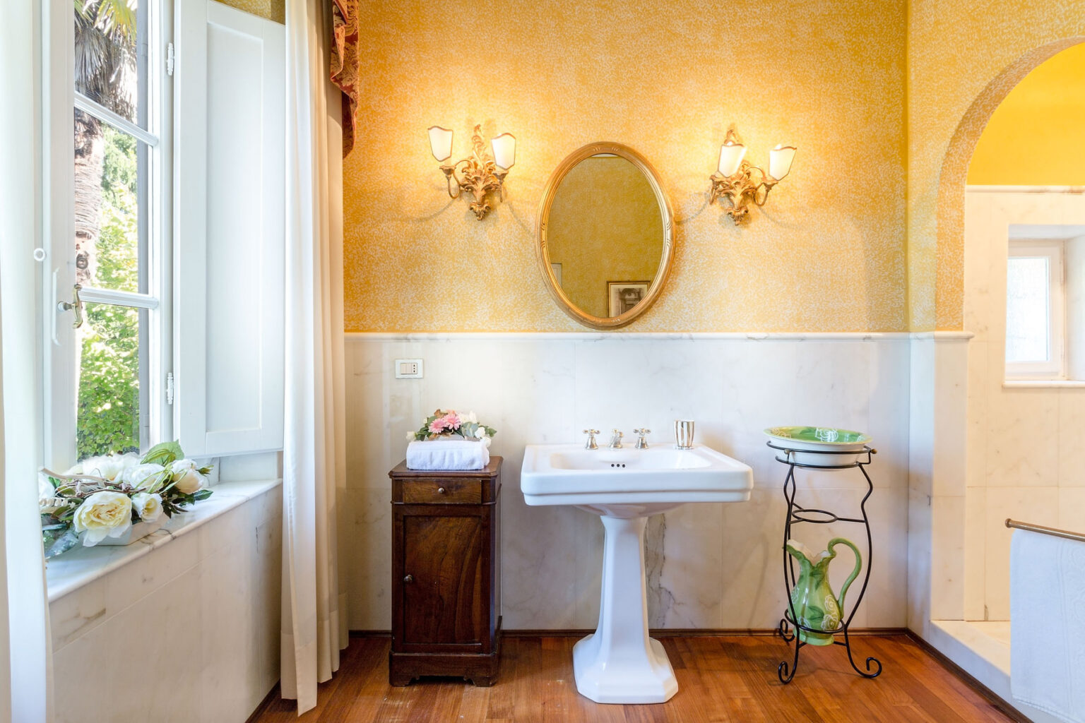 Elegant bathroom interior featuring a vintage sink, ornate mirror, and decorative lighting. The warm yellow walls complement the wooden cabinet and floral arrangements on the windowsill, creating a cozy and inviting atmosphere. | Only on SopranoVillas