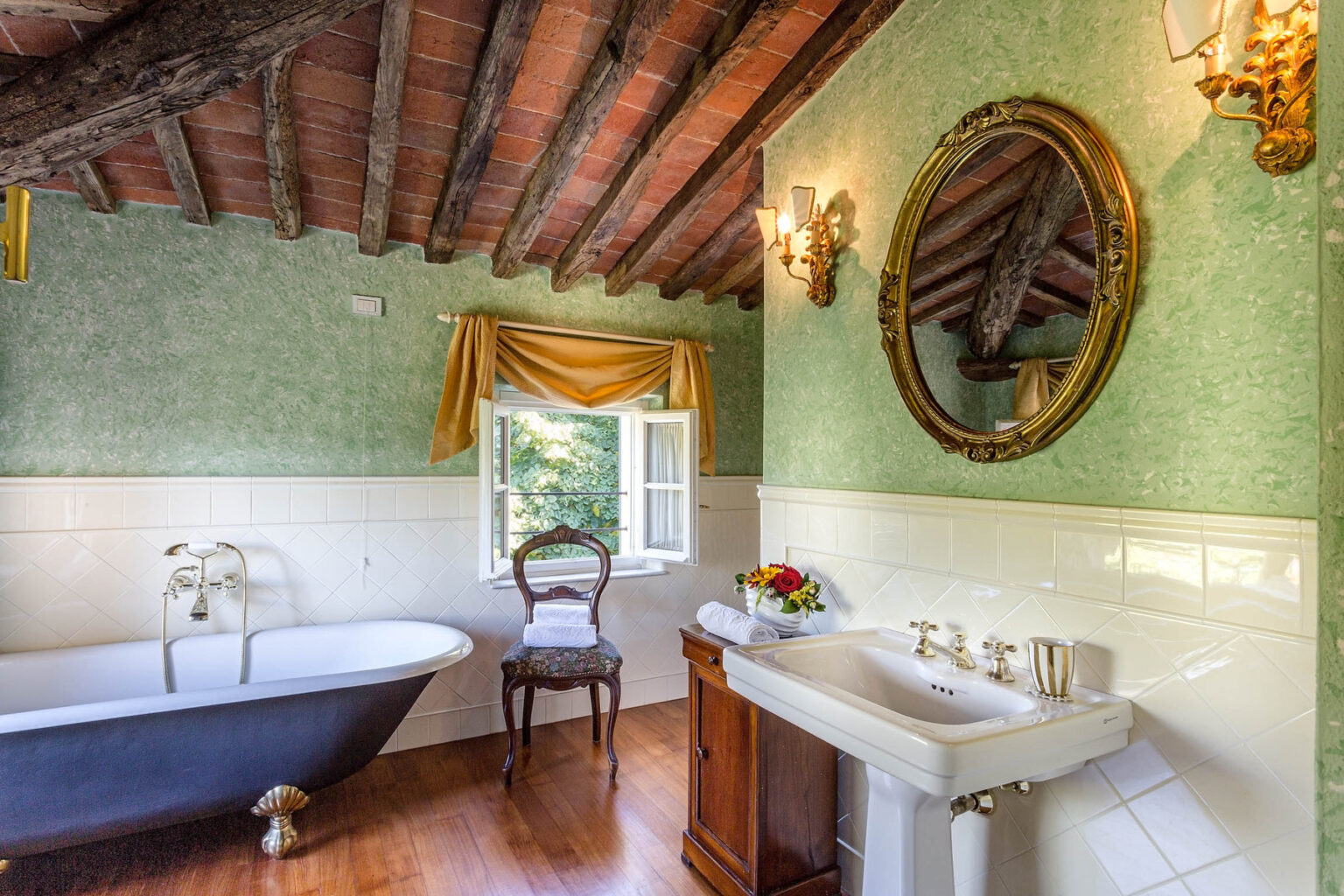 Elegant vintage bathroom featuring a freestanding bathtub, a wooden vanity with a sink, and a decorative mirror. The room has green wallpaper, exposed wooden beams, and a chair next to a window with natural light. Ideal for showcasing classic interior design. | Only on SopranoVillas