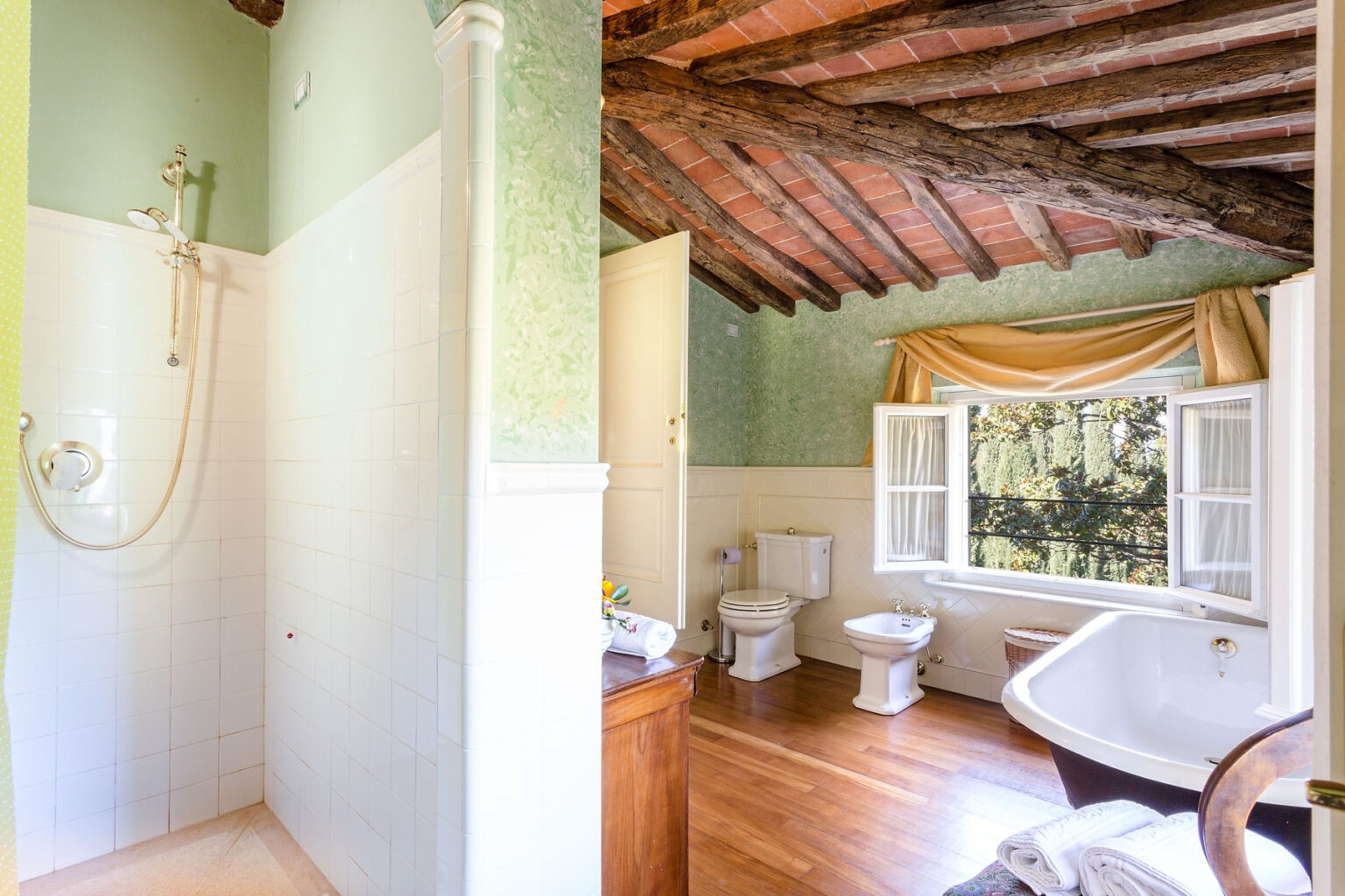 Modern bathroom featuring a shower stall with glass doors and rustic wooden beams. The space includes a toilet, bidet, and a freestanding bathtub beside a large window with elegant drapes, showcasing a bright and airy atmosphere. | Only on SopranoVillas