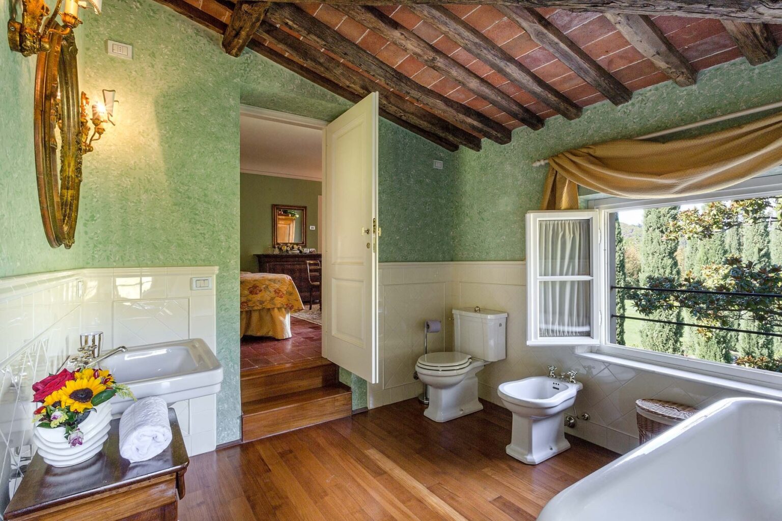Elegant bathroom with rustic wooden beams, featuring a vintage sink, toilet, and bidet. Natural light streams in through a window adorned with soft drapes, complemented by a floral arrangement on a wooden floor. The green wallpaper adds a touch of charm to the serene space. | Only on SopranoVillas