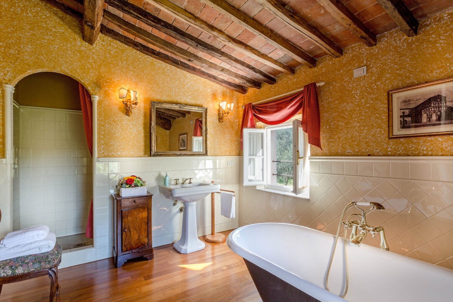 Rustic bathroom featuring a vintage freestanding bathtub, elegant wooden beams, and warm yellow walls. The space includes a pedestal sink, a shower area, and decorative accents like a framed picture and floral arrangement, creating a cozy and inviting atmosphere. Natural light floods in through the window, enhancing the charming ambiance. | Only on SopranoVillas