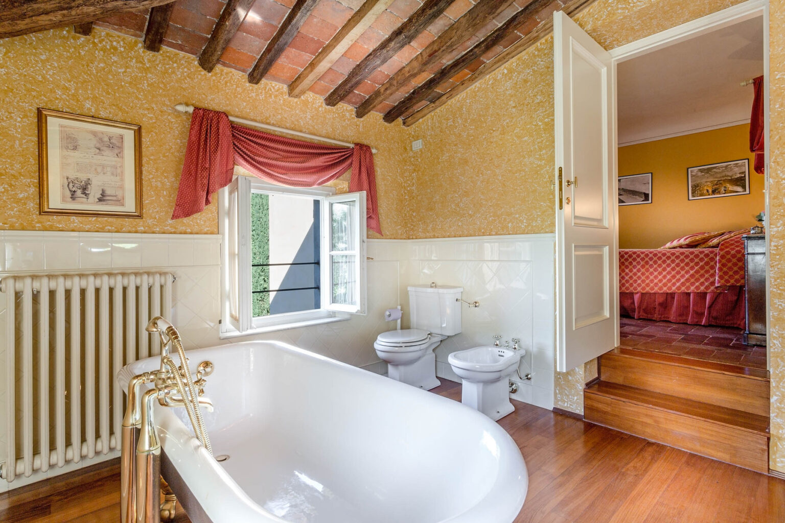 Spacious bathroom featuring a classic freestanding bathtub, elegant wooden beams, and a cozy atmosphere. The room includes a radiator, modern toilet, and bidet, with a view of a well-decorated bedroom through an open doorway. | Only on SopranoVillas