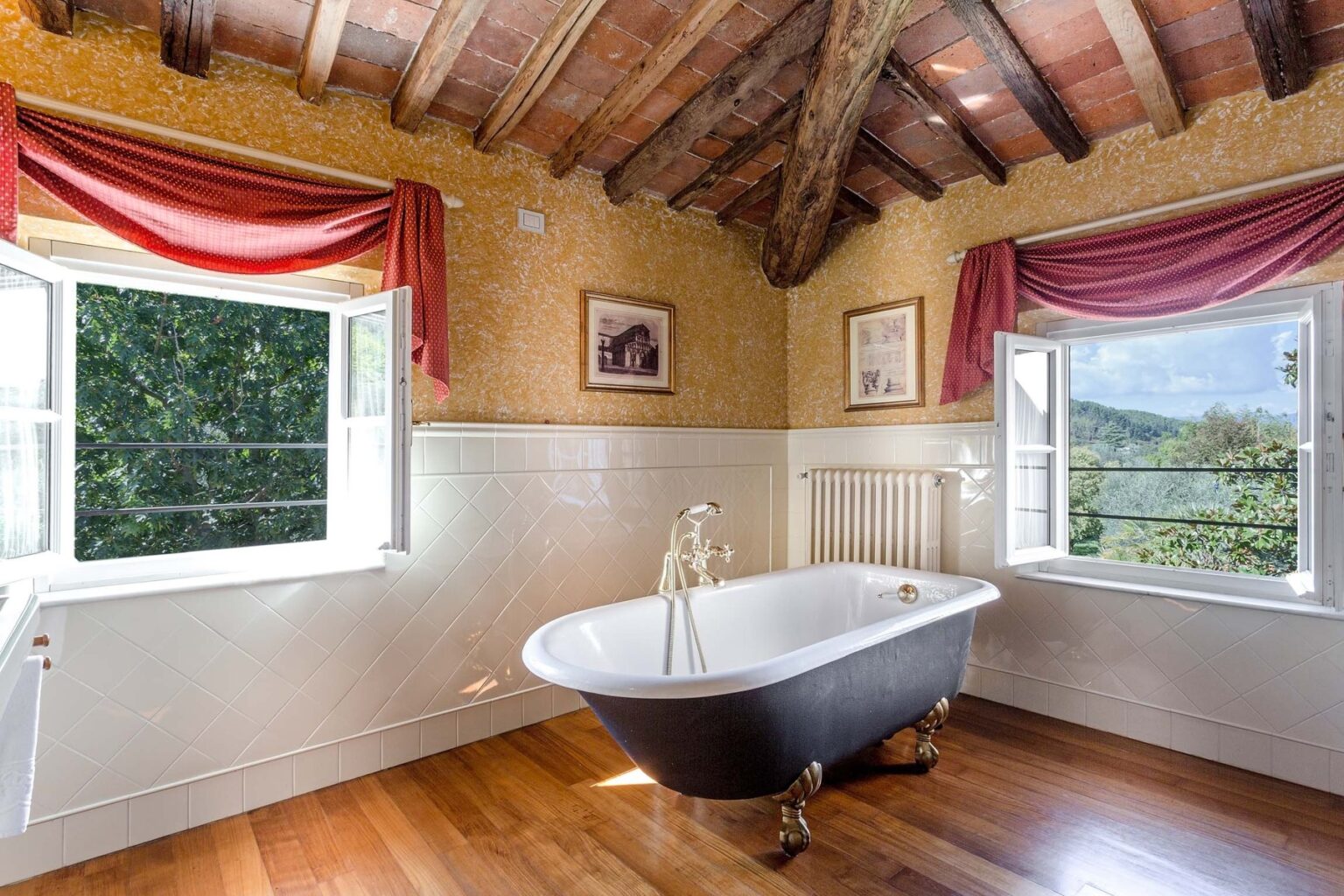 Elegant bathroom featuring a vintage freestanding bathtub, wooden beams, and large windows with scenic views. The warm, inviting decor includes rich wooden flooring and colorful drapes, creating a cozy atmosphere. Perfect for a relaxing retreat. | Only on SopranoVillas
