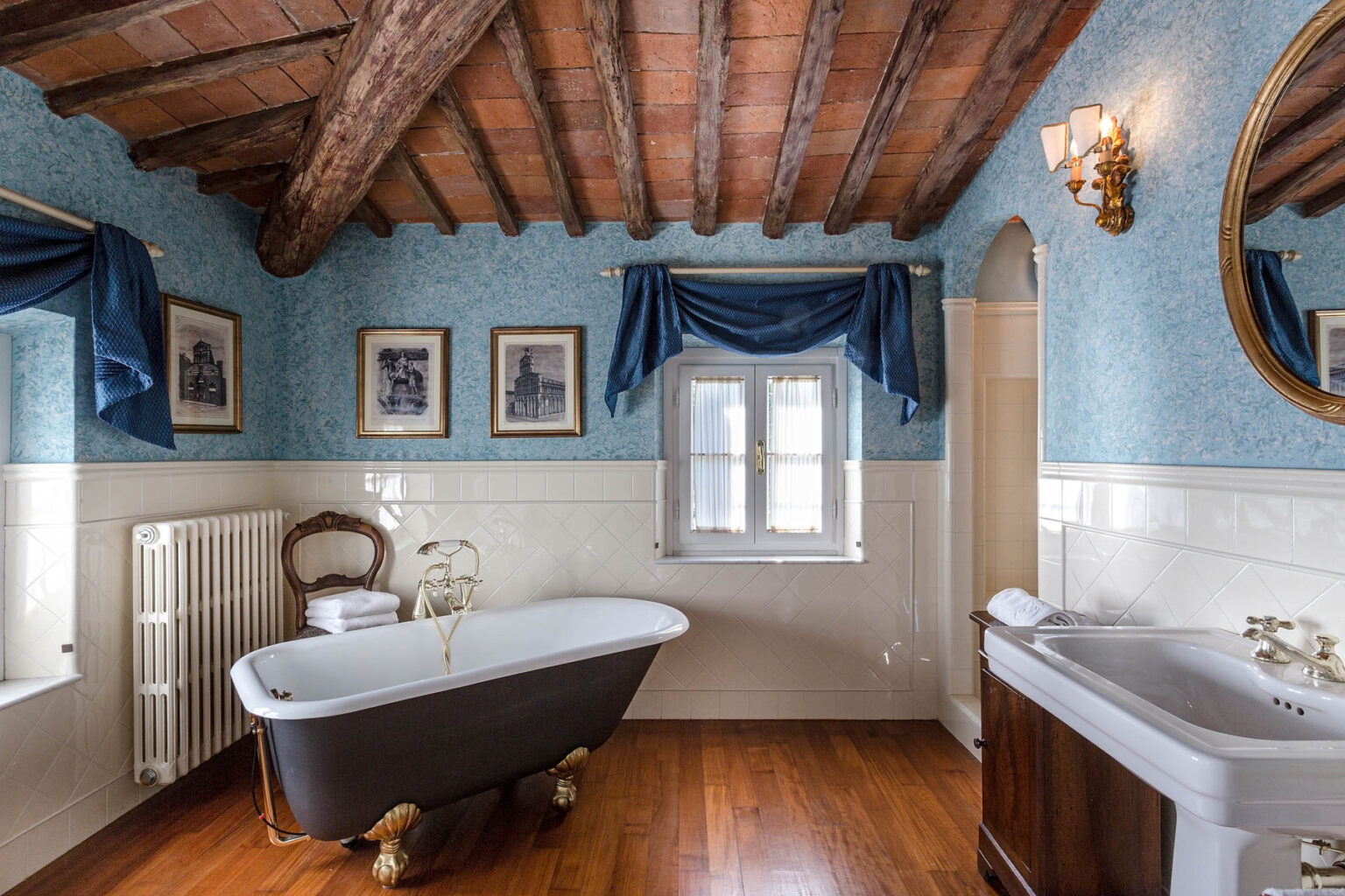 Elegant bathroom featuring a vintage freestanding bathtub with gold accents, wooden beams on the ceiling, blue floral wallpaper, and framed artwork. The space includes a classic wooden chair, a white radiator, and a stylish vanity, creating a cozy and inviting atmosphere. | Only on SopranoVillas