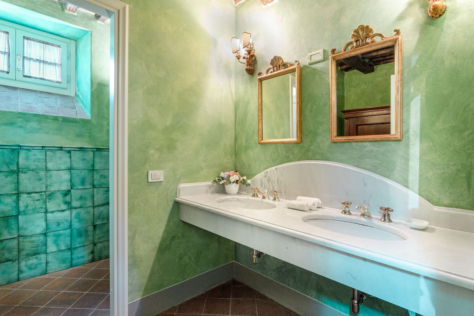 Elegant bathroom with green textured walls, featuring a white double sink countertop, vintage mirrors, and decorative lighting. The space is complemented by a stylish tile backsplash and natural light from a nearby window. | Only on SopranoVillas