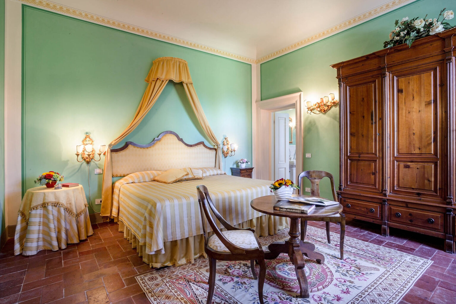 Elegant bedroom featuring mint green walls, a canopied bed with striped bedding, antique wooden furniture, and a cozy seating area with a round table and floral decor. | Only on SopranoVillas