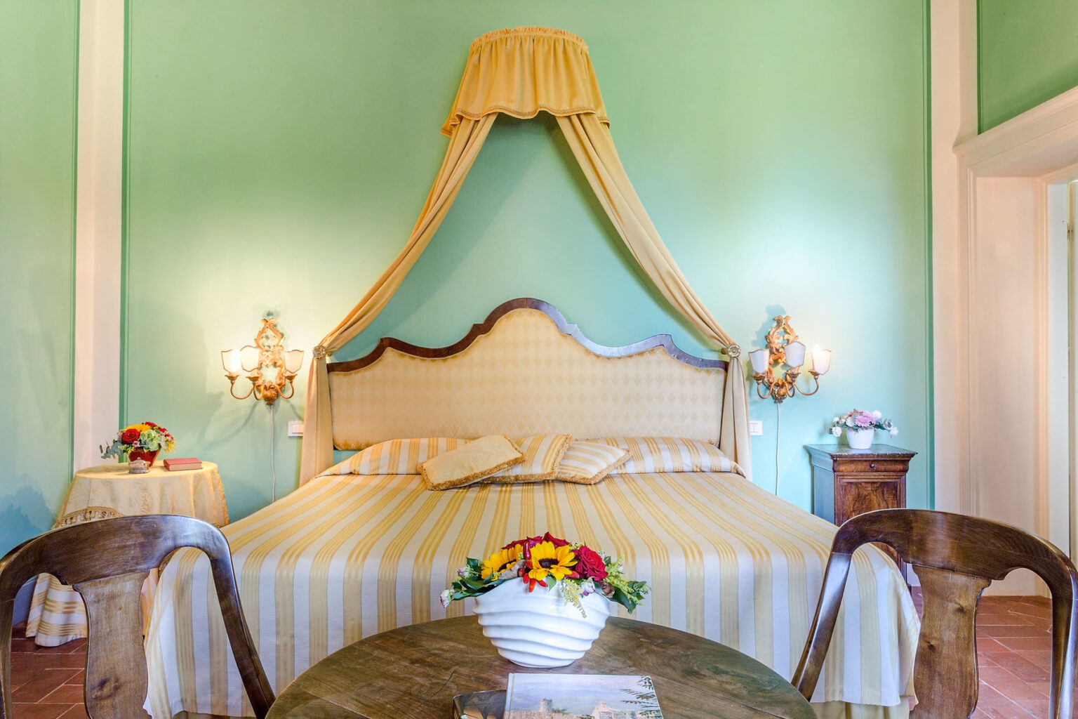 Elegant bedroom featuring a green wall, a striped yellow bedspread, and a decorative canopy above the bed. Two bedside tables with floral arrangements and a small round table with a vase of flowers complete the inviting ambiance. | Only on SopranoVillas