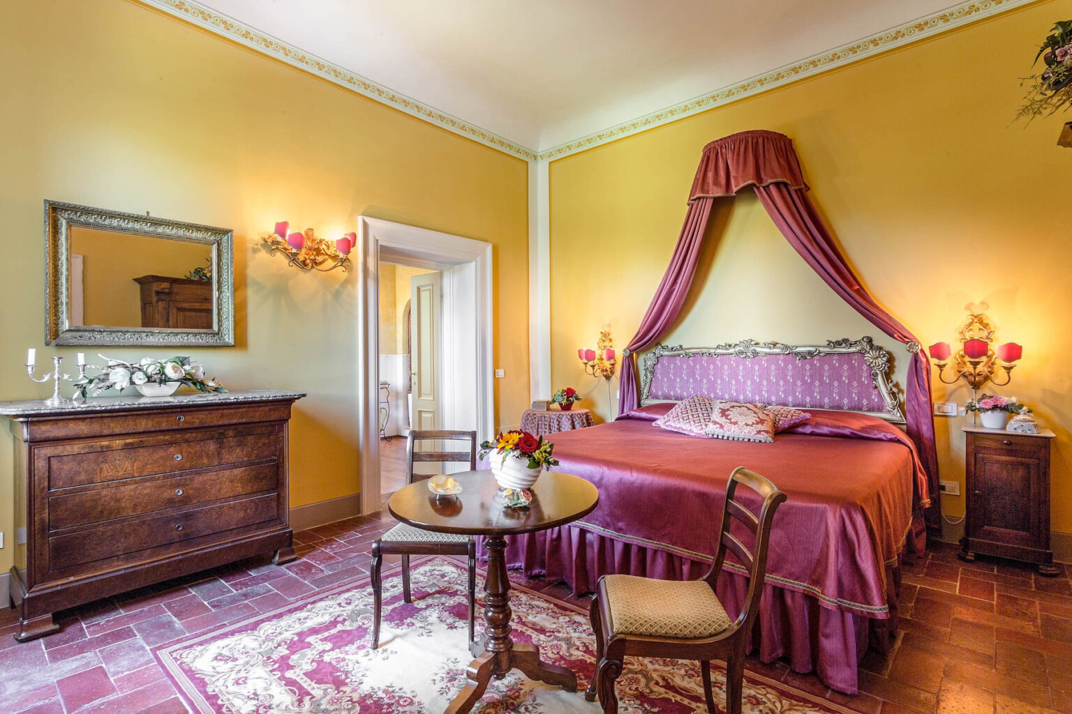 Elegant bedroom featuring a yellow wall, a luxurious bed with a canopy, vintage furniture including a wooden dresser, and a cozy seating area with a small table and floral decor, perfect for a relaxing getaway. | Only on SopranoVillas