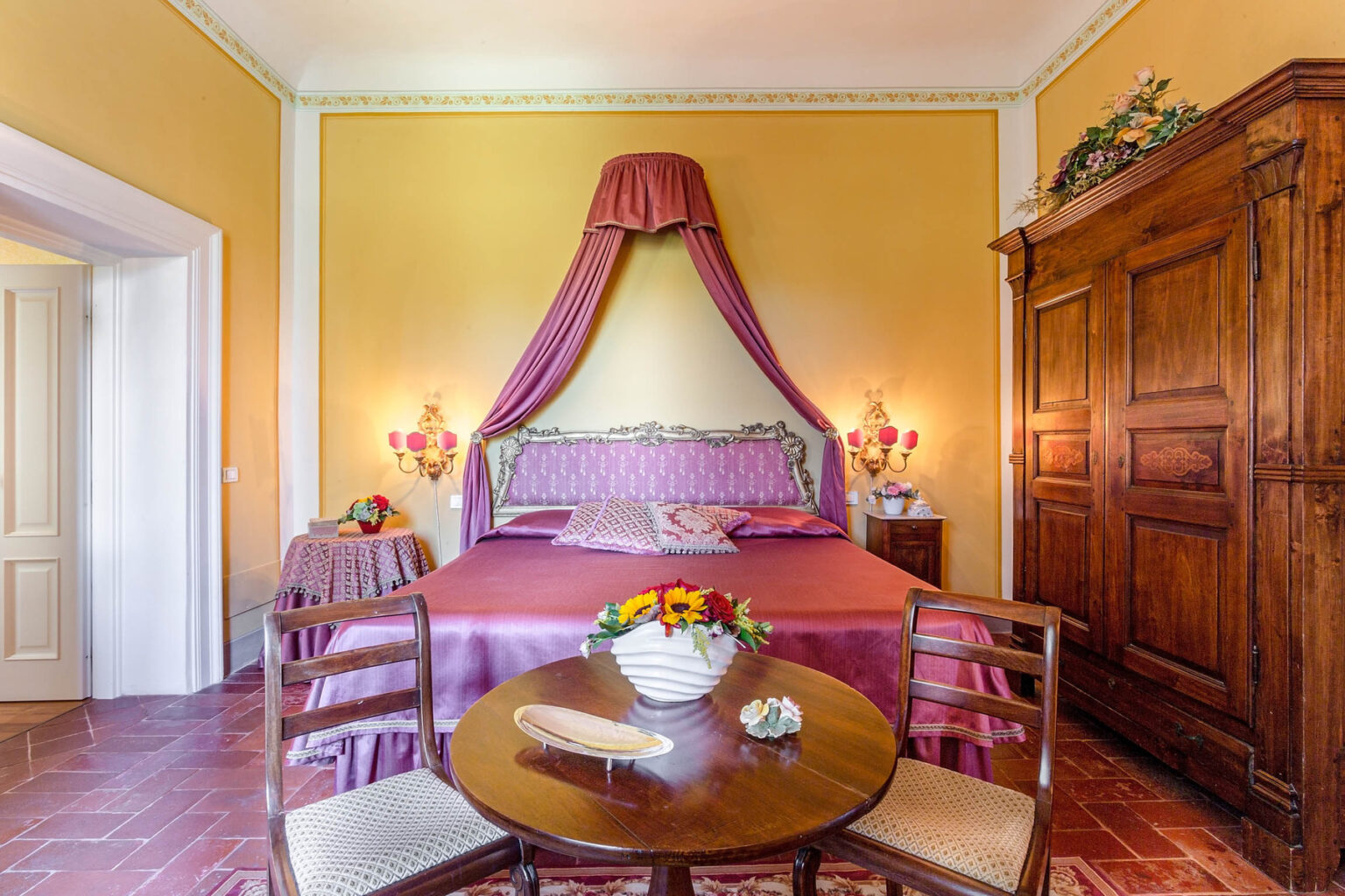 Elegant bedroom with warm yellow walls, a plush pink bed adorned with a canopy, and a wooden wardrobe. A round table with a decorative vase of flowers adds charm to the inviting space. | Only on SopranoVillas