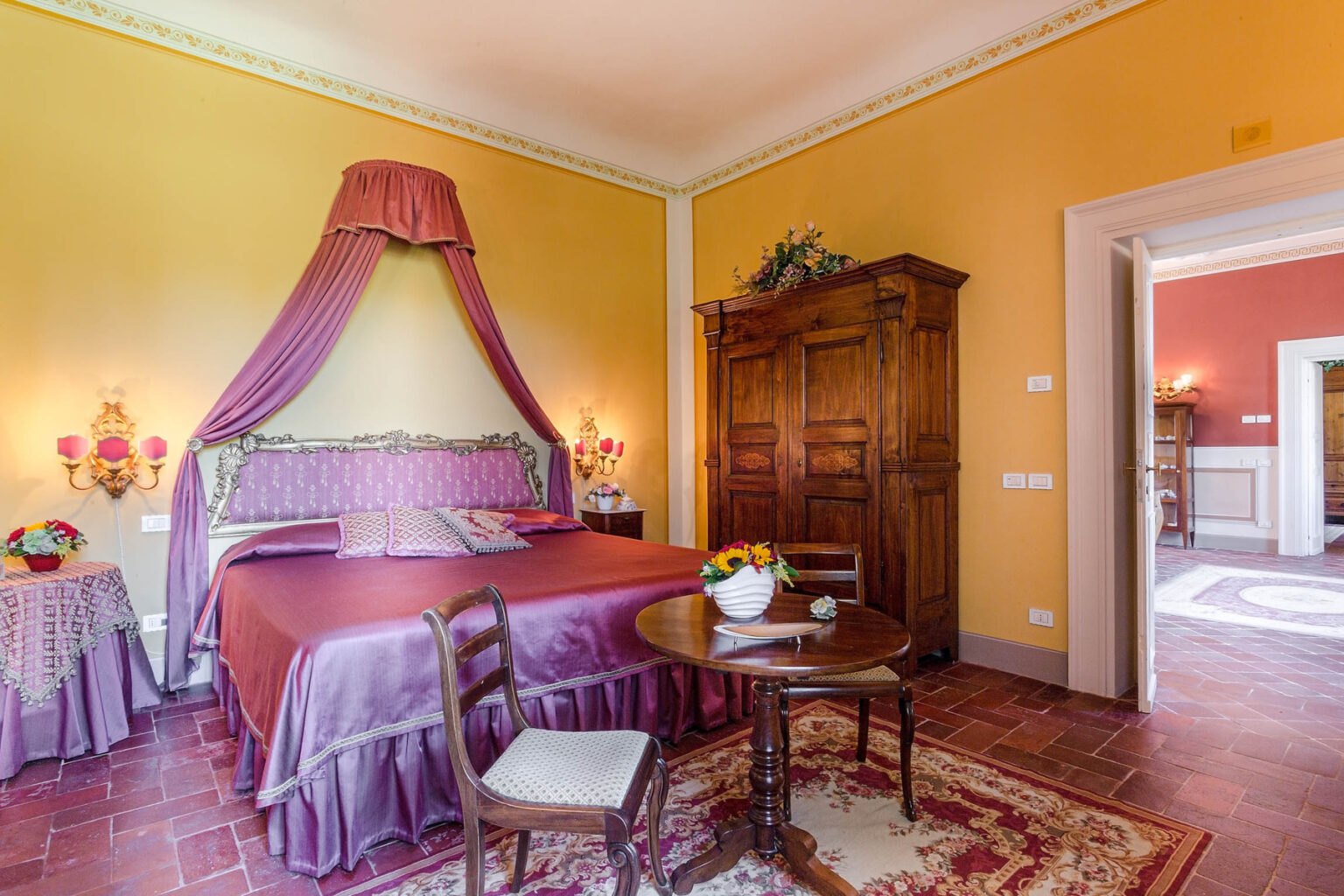Elegant bedroom featuring a canopy bed draped in purple fabric, vintage wooden wardrobe, and a round table adorned with a fruit bowl, set against warm yellow walls and classic decor. | Only on SopranoVillas