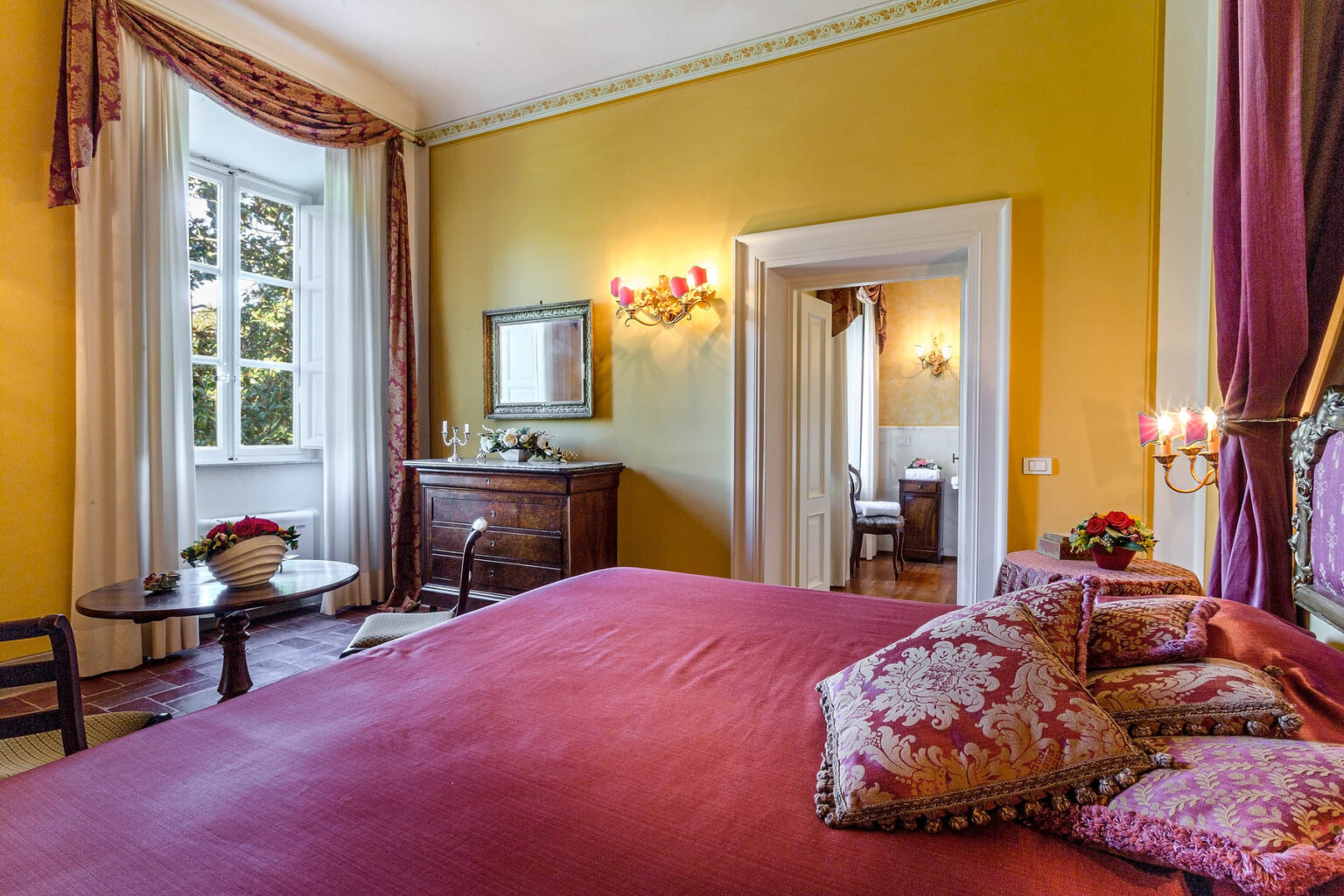 Cozy and elegantly decorated bedroom featuring a vibrant yellow wall, a large red bedspread, ornate pillows, and classic furnishings, with natural light streaming through a window. | Only on SopranoVillas