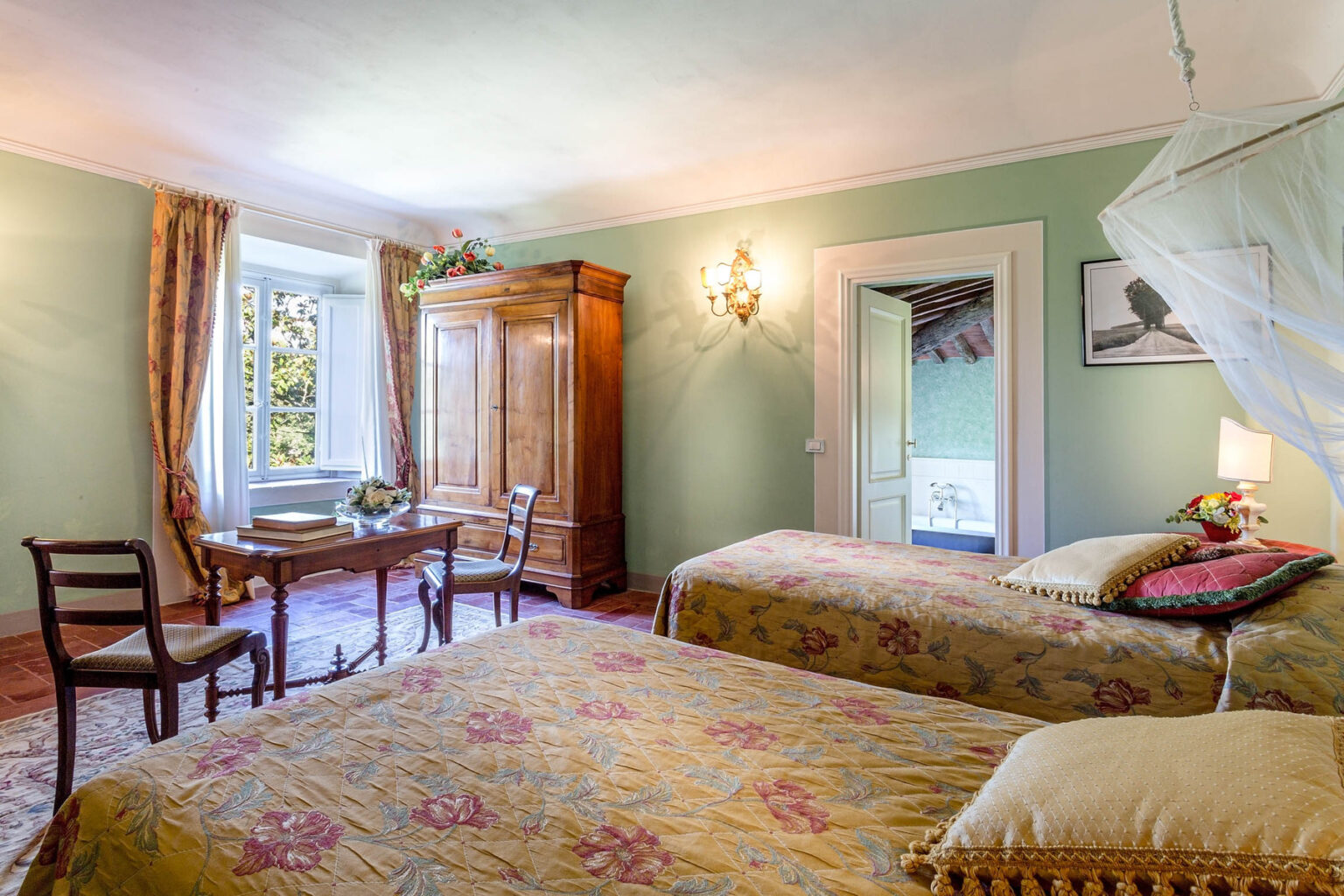 Cozy guest room featuring two beds with floral bedding, a wooden wardrobe, and a small dining table. The room is adorned with soft green walls and elegant curtains, providing a warm and inviting atmosphere. Natural light streams in through the window, enhancing the serene ambiance. | Only on SopranoVillas