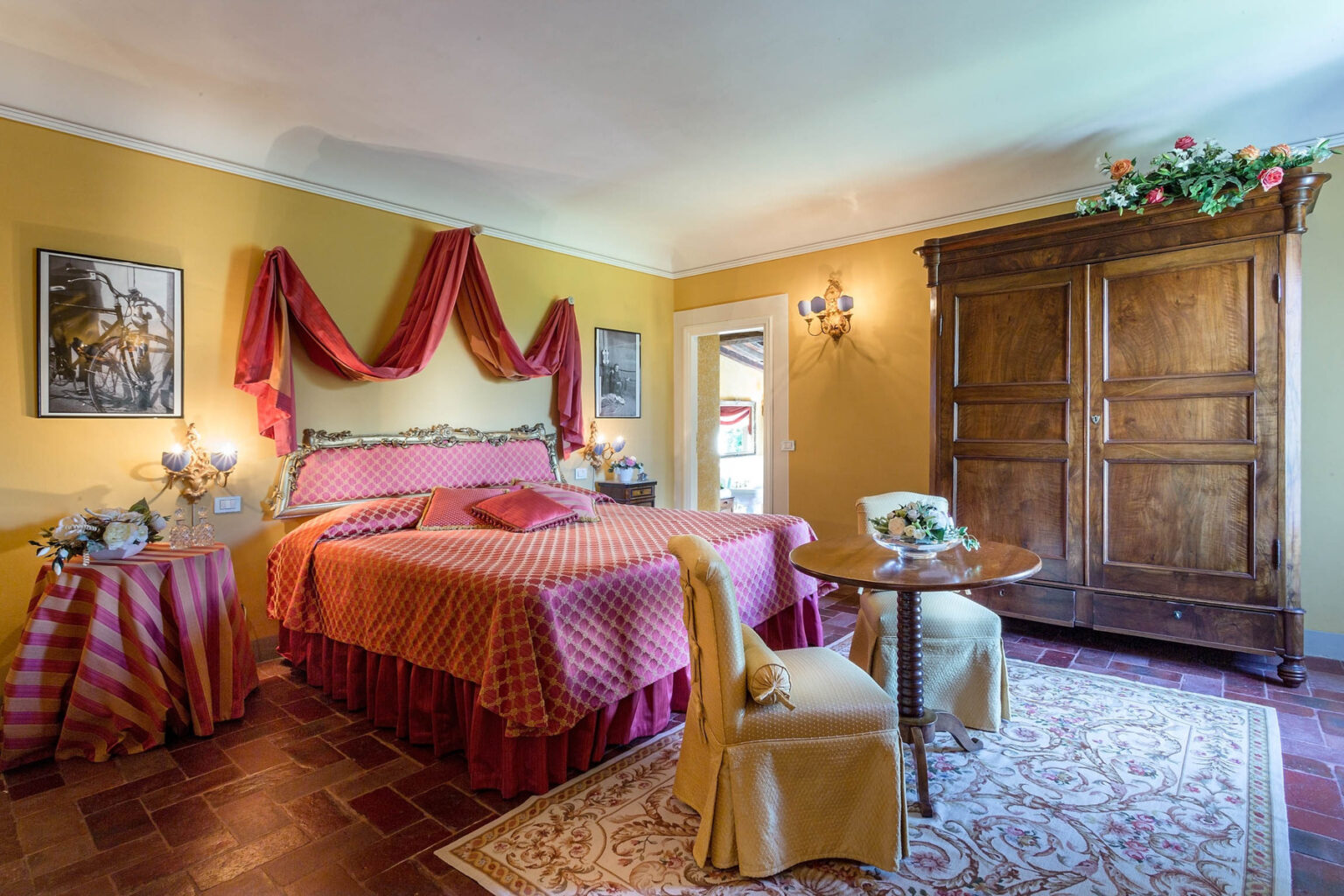 Cozy and elegantly decorated hotel room featuring a king-sized bed with a red patterned cover, warm yellow walls, and classic wooden furniture. The space includes a small dining table and a vintage wardrobe, creating a welcoming atmosphere for guests. | Only on SopranoVillas