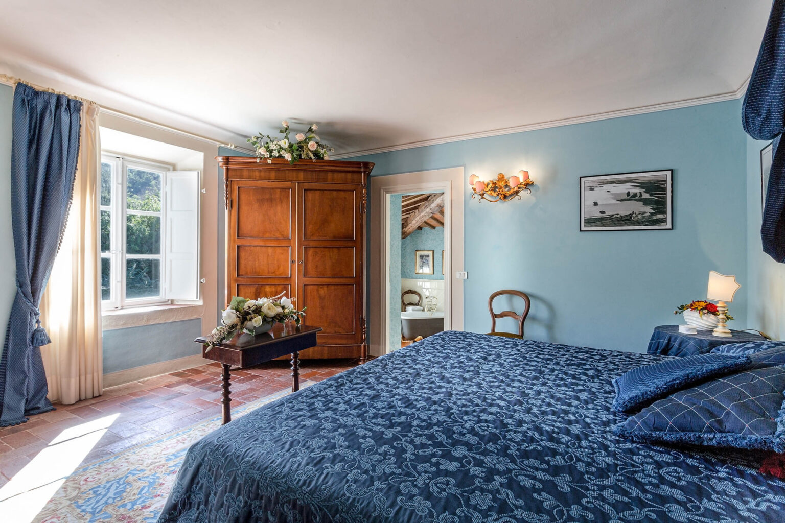 Cozy bedroom featuring blue walls, a queen-sized bed with a blue patterned duvet, wooden furniture, and large windows allowing natural light. The room includes a wardrobe, a decorative chair, and floral accents, creating a serene and inviting atmosphere. | Only on SopranoVillas