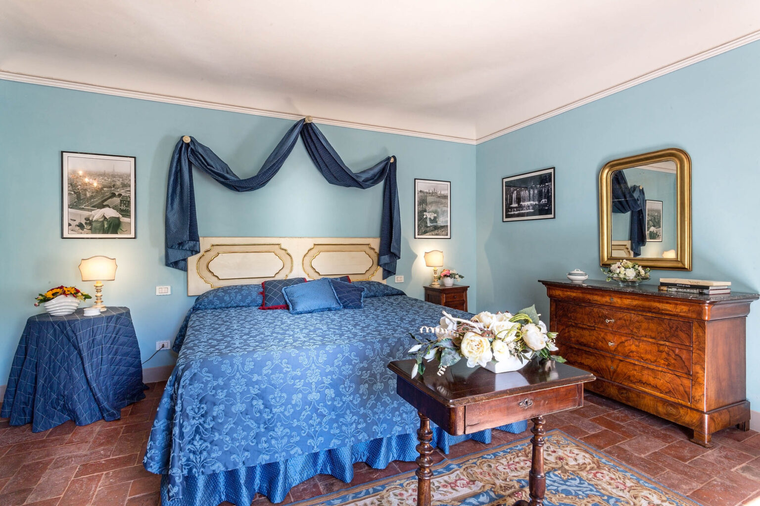 Cozy bedroom featuring a blue color scheme, a large bed with decorative pillows, vintage furniture, and elegant lighting. The walls are painted light blue, complemented by framed artwork and a stylish mirror, creating a welcoming atmosphere. | Only on SopranoVillas
