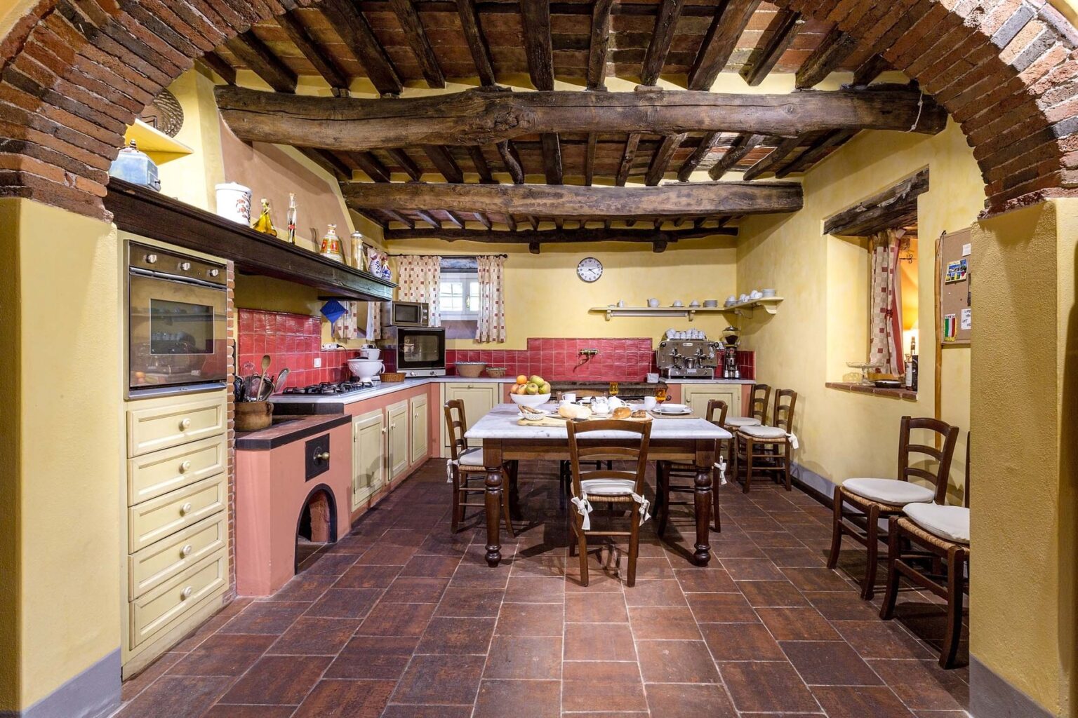 Rustic kitchen interior featuring exposed wooden beams, a large dining table with wooden chairs, and a colorful backsplash. The space is warmly lit, creating an inviting atmosphere for cooking and dining. | Only on SopranoVillas