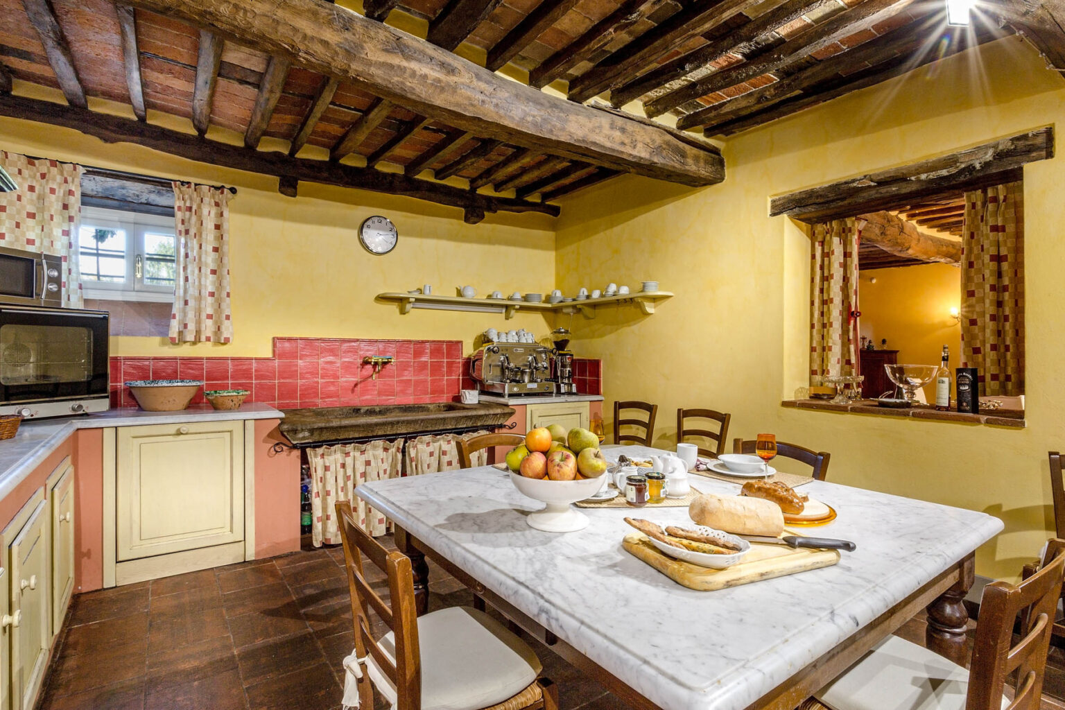 Rustic kitchen interior featuring wooden beams, a large dining table with bread and fruit, yellow walls, and vintage decor. The space includes a microwave, oven, and shelves with kitchenware, creating a cozy and inviting atmosphere. | Only on SopranoVillas