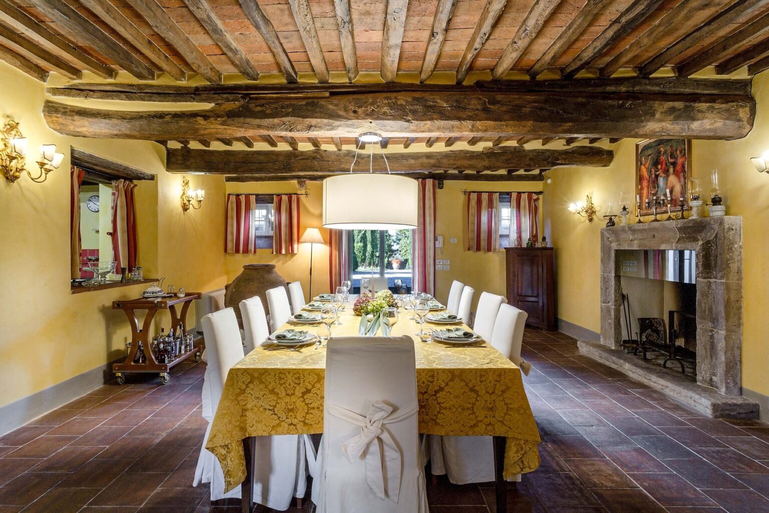 Spacious dining room with rustic wooden beams, a large table set for a meal, and a cozy fireplace, featuring warm yellow walls and natural light from windows. | Only on SopranoVillas