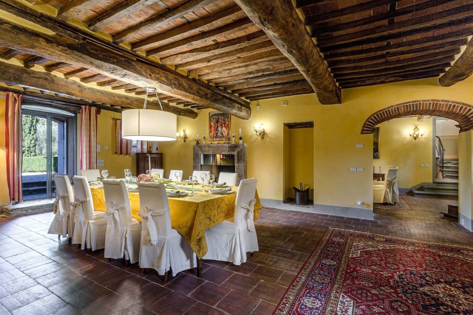 Elegant dining room featuring a rustic wooden beam ceiling, a large table set with fine dining ware, and warm yellow walls. The space is illuminated by a stylish chandelier and includes large windows with scenic views. A cozy atmosphere enhanced by decorative elements and a plush area rug. | Only on SopranoVillas