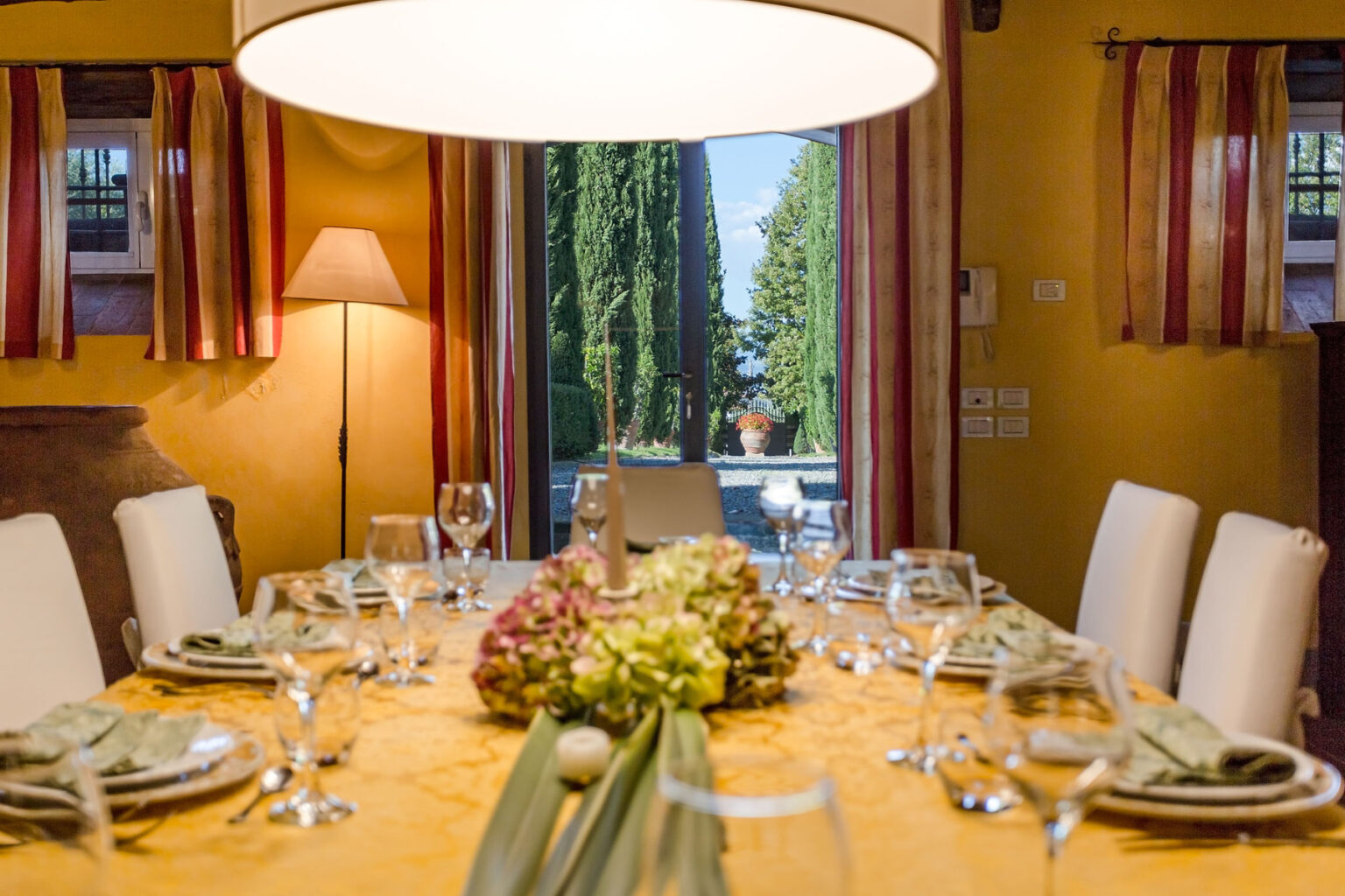 Elegant dining room setup featuring a long table adorned with glassware and a floral centerpiece, surrounded by bright yellow walls and large windows showcasing a garden view. | Only on SopranoVillas