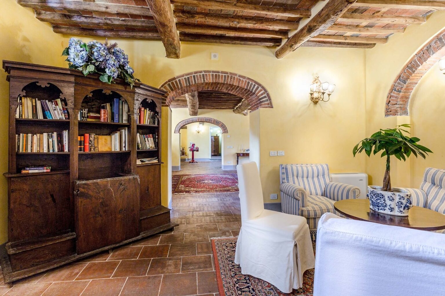 Cozy living room featuring a wooden bookshelf filled with books, comfortable seating, and warm yellow walls, complemented by rustic wooden beams and elegant lighting. | Only on SopranoVillas