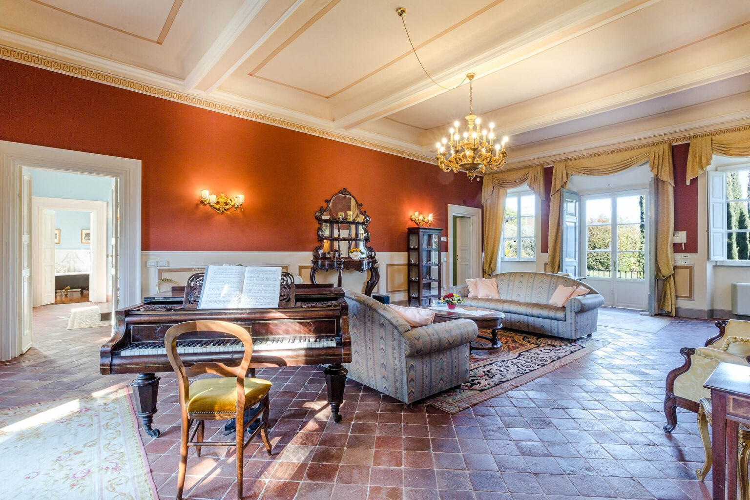 Elegant living room featuring a grand piano, vintage furnishings, and large windows allowing natural light, with a rich red accent wall and ornate chandelier. | Only on SopranoVillas