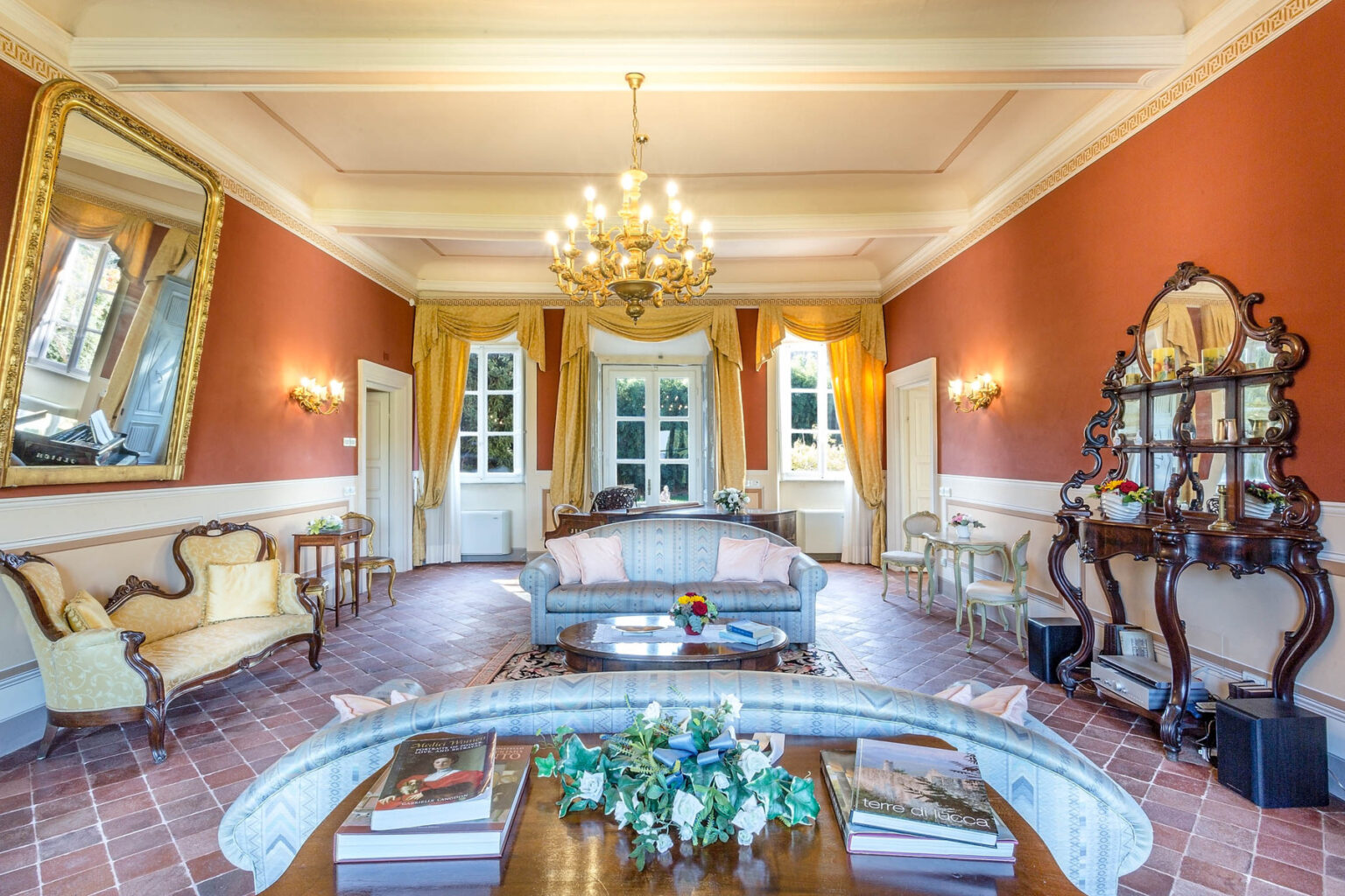 Elegant living room featuring a vintage chandelier, ornate mirror, and classic furniture. Warm orange walls complement the natural light streaming through large windows. A decorative coffee table with flowers adds a touch of sophistication. | Only on SopranoVillas