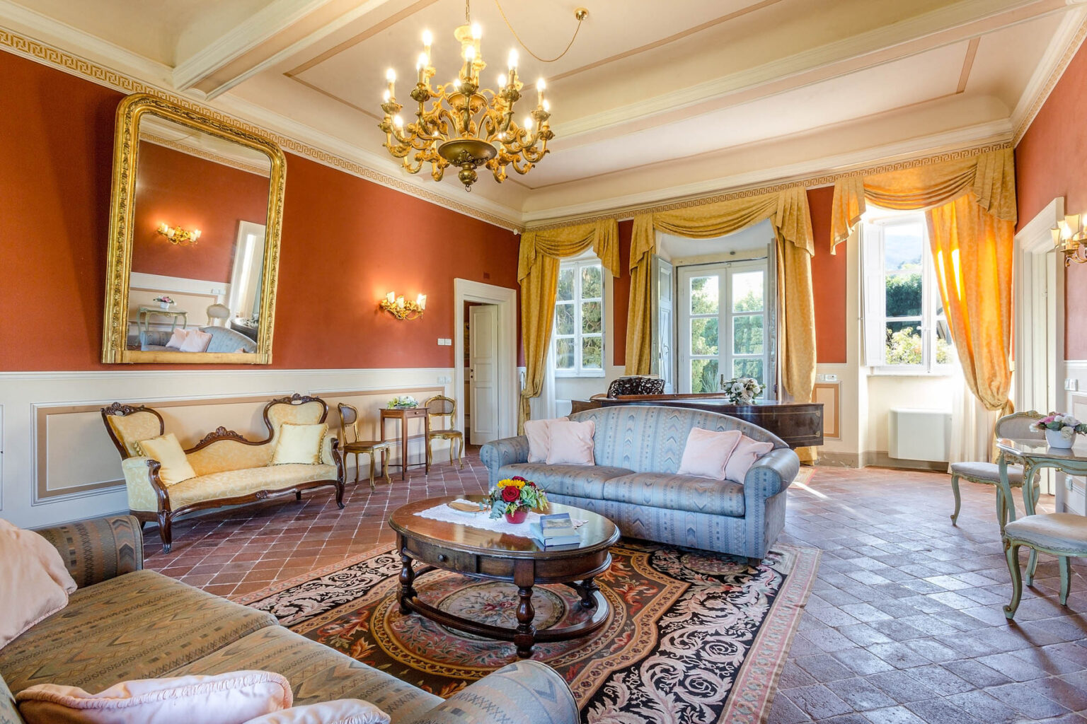 Elegant living room interior featuring vintage furniture, a large chandelier, and warm orange walls. Natural light streams through large windows, illuminating a cozy seating area with a coffee table and decorative flowers. Ideal for showcasing luxurious home design. | Only on SopranoVillas