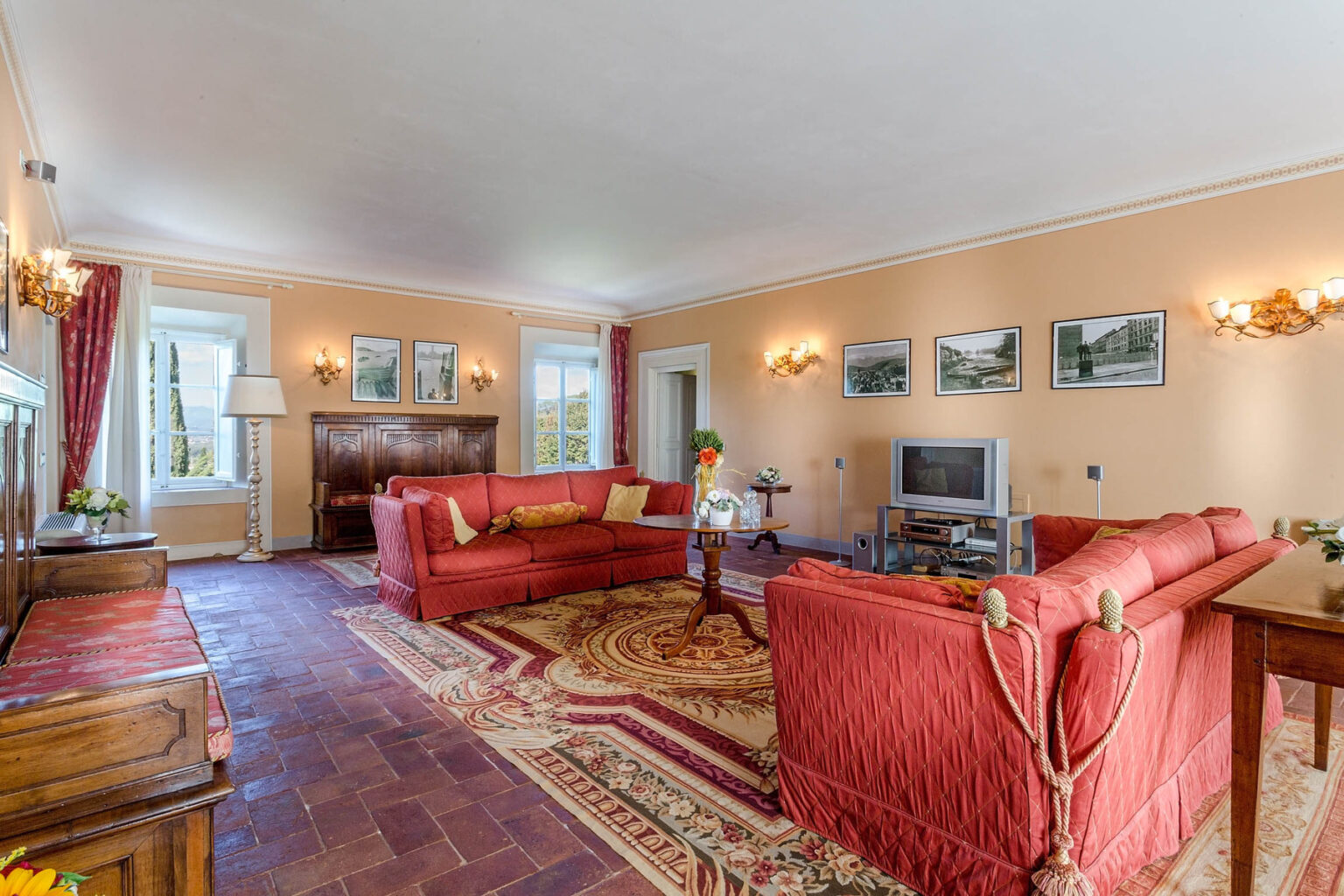 Spacious living room featuring red sofas, a decorative area rug, wooden furniture, and large windows that provide natural light, complemented by framed landscape photographs on the walls. | Only on SopranoVillas