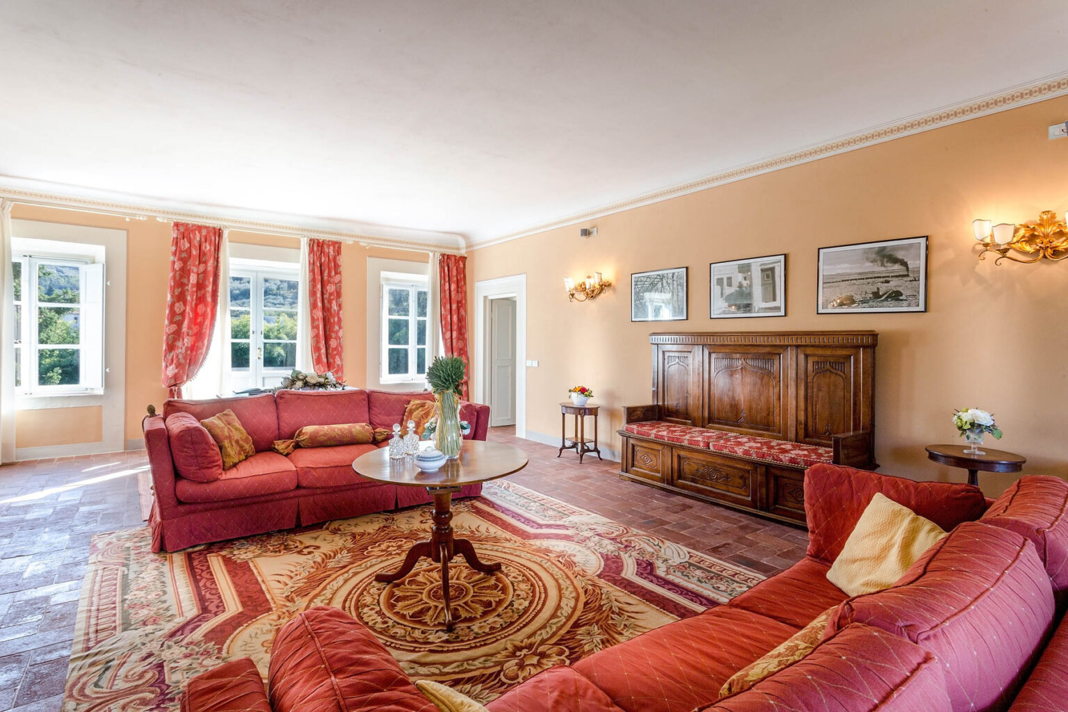 Spacious living room featuring elegant red sofas, a round coffee table, and decorative wall art, illuminated by natural light from large windows with patterned curtains. | Only on SopranoVillas