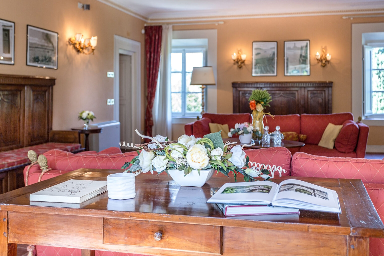 Cozy living room interior featuring a wooden table adorned with flowers, a decorative vase, and books. The background includes a red sofa, elegant lighting, and framed artwork on the walls, creating a warm and inviting atmosphere. | Only on SopranoVillas