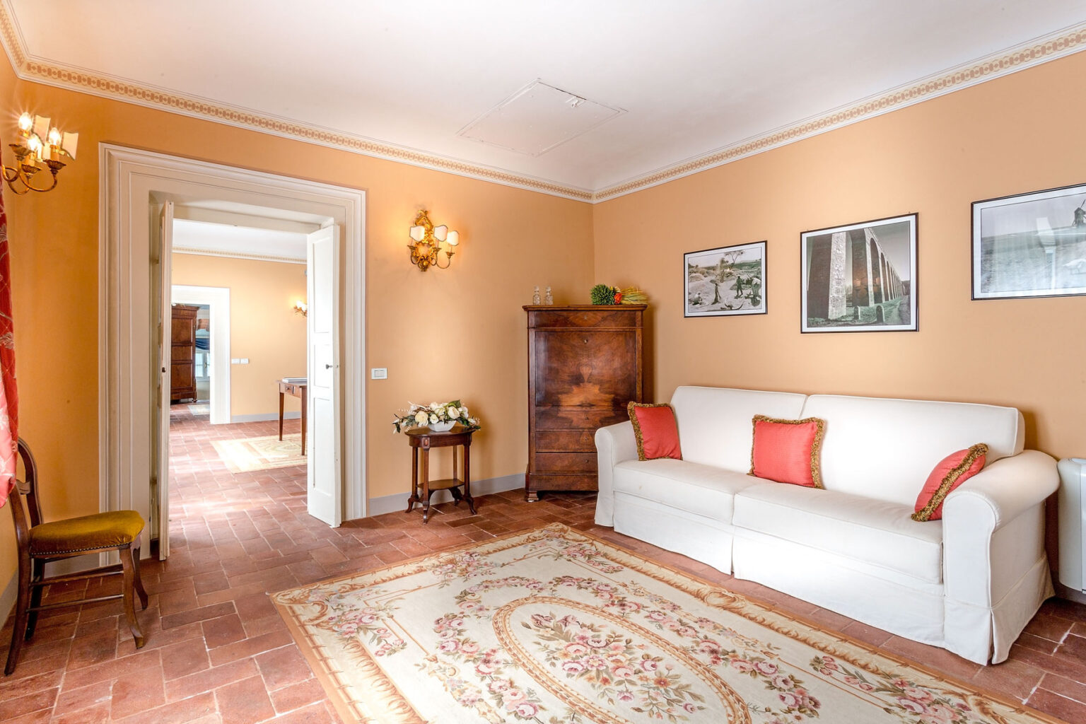Cozy living room featuring a white sofa with red cushions, a vintage wooden cabinet, and framed artwork on the orange walls. The room has a patterned rug on a terracotta tile floor, illuminated by wall sconces, creating a warm and inviting atmosphere. | Only on SopranoVillas