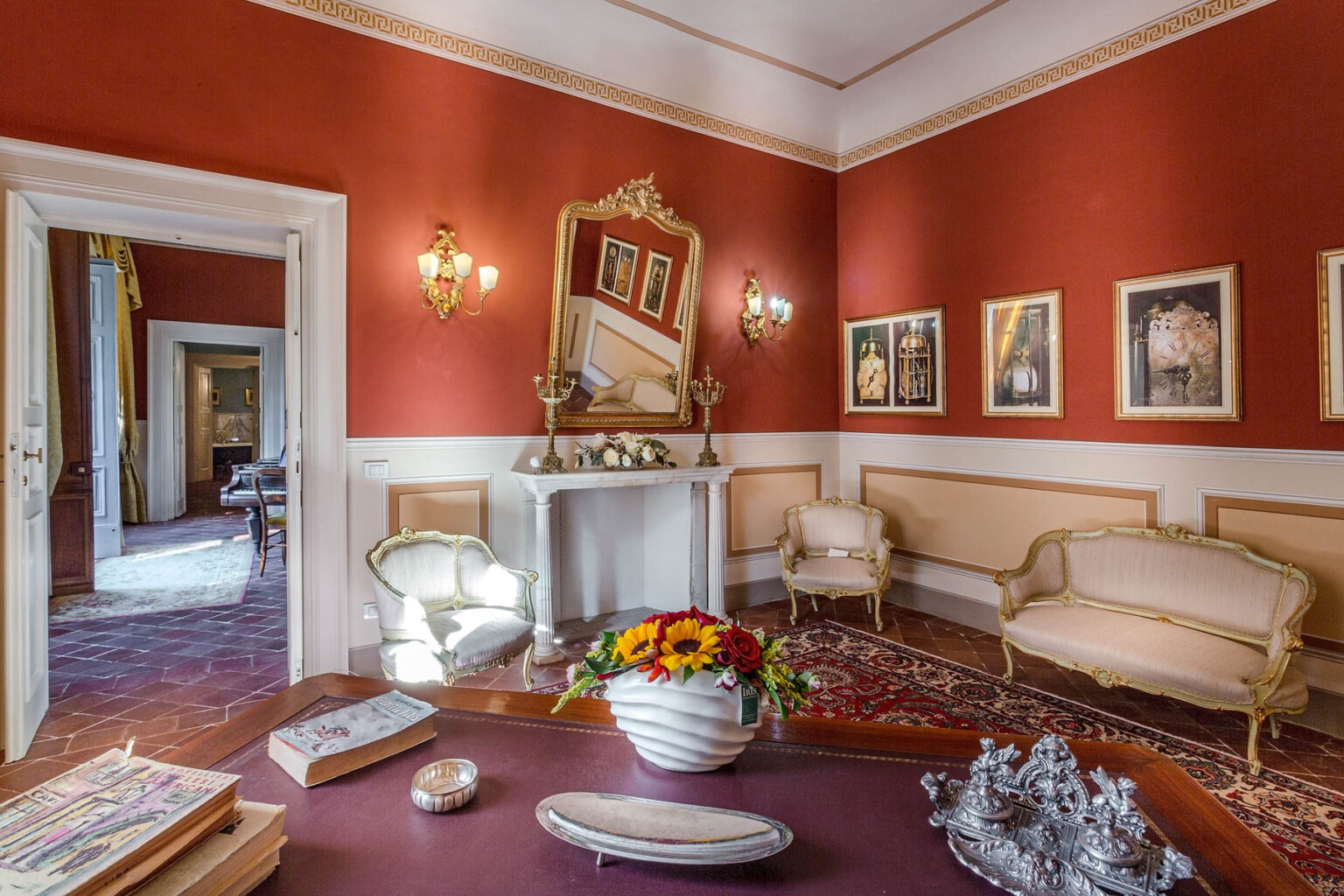 Elegant living room featuring red walls, vintage furniture, and decorative art. The space includes ornate gold-framed mirrors, a floral centerpiece, and warm lighting, creating a sophisticated atmosphere. A glimpse of adjacent rooms is visible through open doors. | Only on SopranoVillas