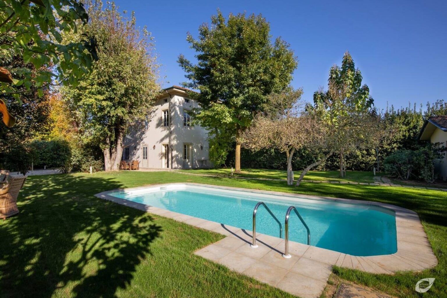 A sunny backyard featuring a sparkling swimming pool surrounded by lush greenery and trees, with a modern house in the background. Ideal for outdoor relaxation and entertaining. | Only on SopranoVillas