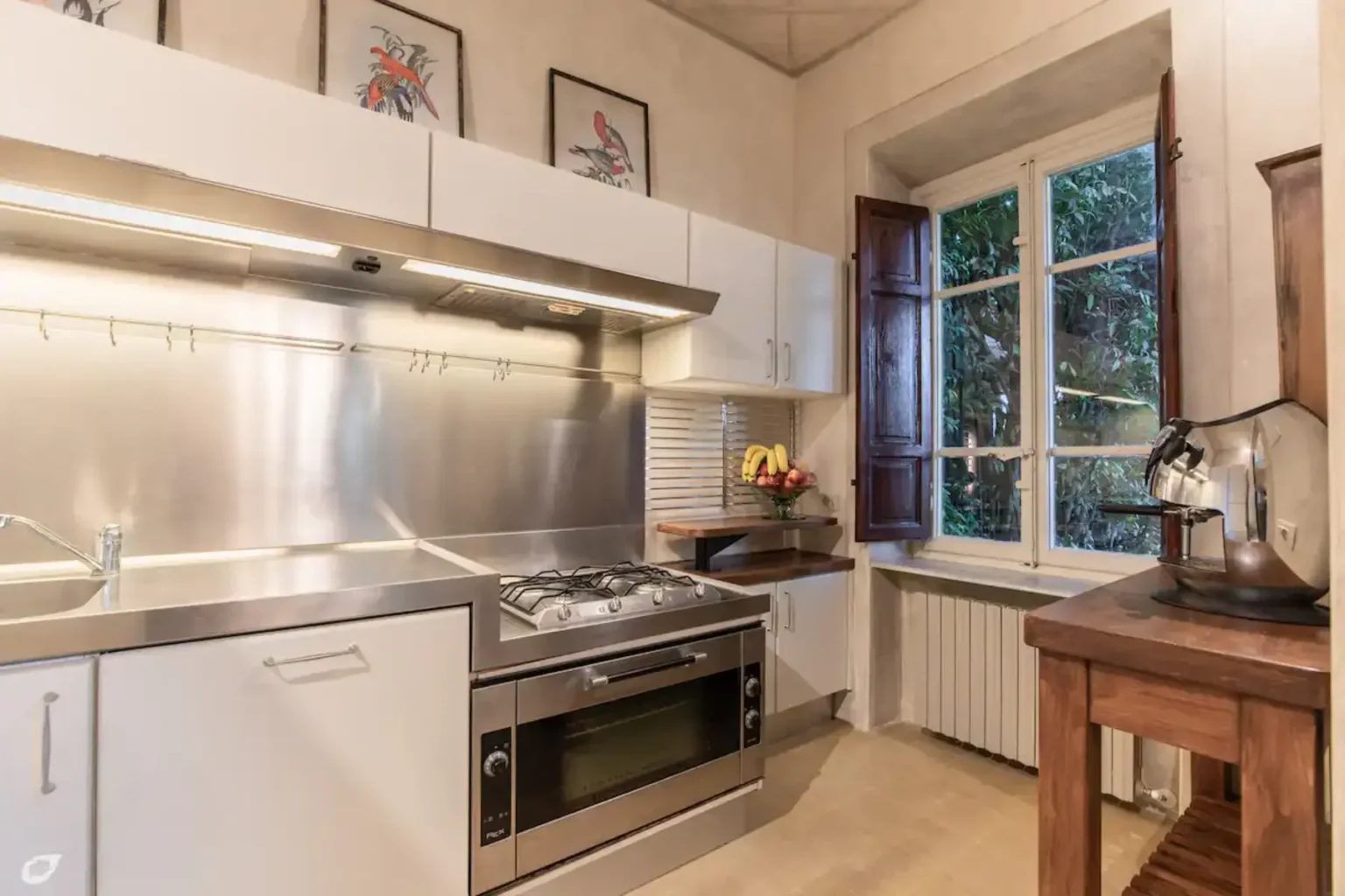 Modern kitchen with stainless steel appliances, white cabinetry, and a window providing natural light. Features a gas stove, wooden table, and decorative artwork on the walls. | Only on SopranoVillas