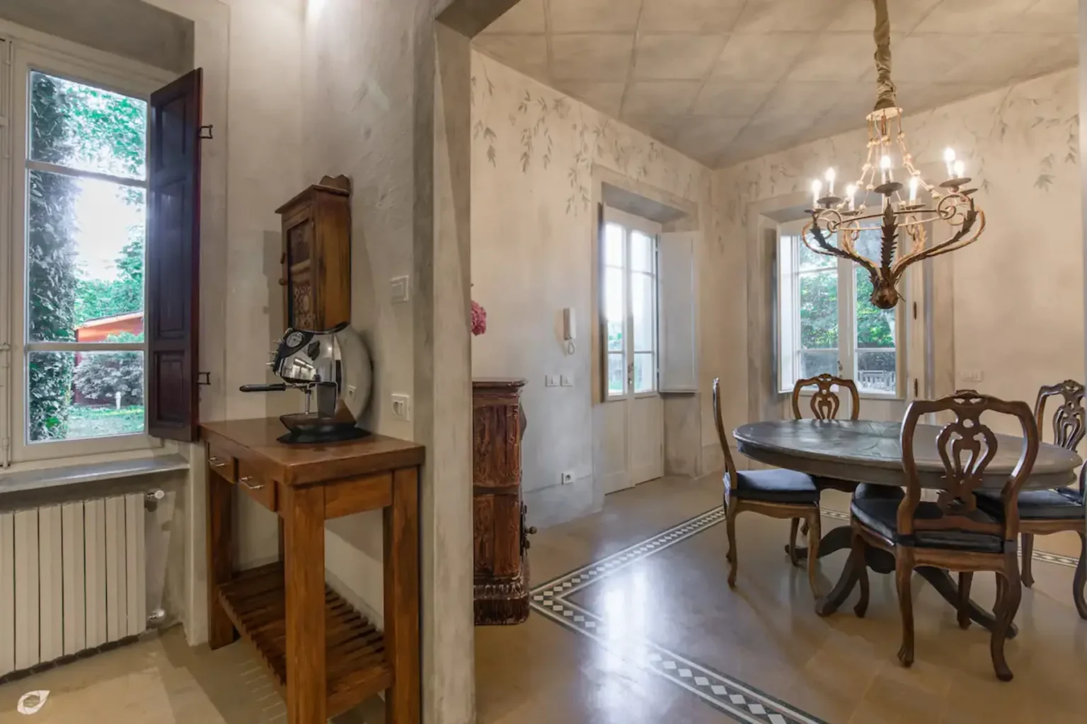 Cozy dining area featuring a wooden table with elegant chairs, a vintage chandelier, and a rustic side table with a coffee machine. Natural light streams in through large windows, highlighting the charming interior design and serene outdoor view. | Only on SopranoVillas