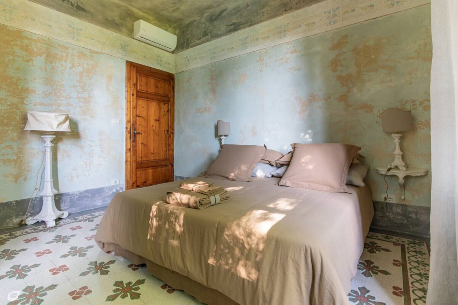 Cozy bedroom featuring a neutral color palette, a comfortable double bed with beige linens, vintage bedside lamps, and a wooden door. The room has textured walls and patterned tile flooring, creating a warm and inviting atmosphere. | Only on SopranoVillas