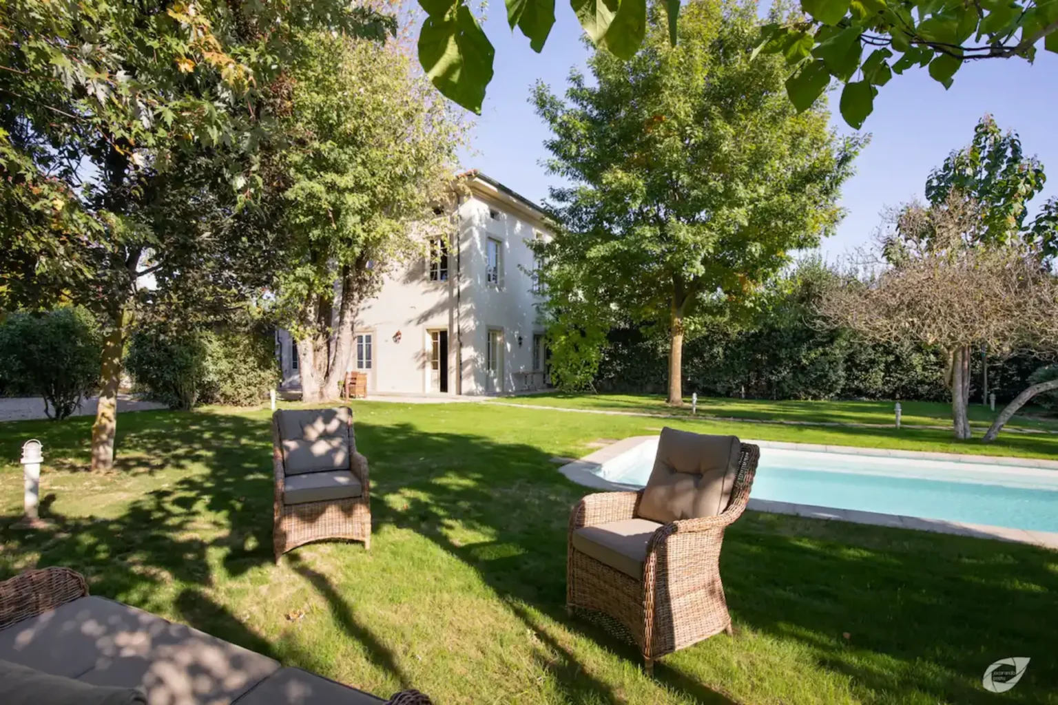 Charming backyard with a well-maintained lawn, two wicker chairs, and a swimming pool, surrounded by trees and a modern house in the background. Perfect for outdoor relaxation and entertaining. | Only on SopranoVillas
