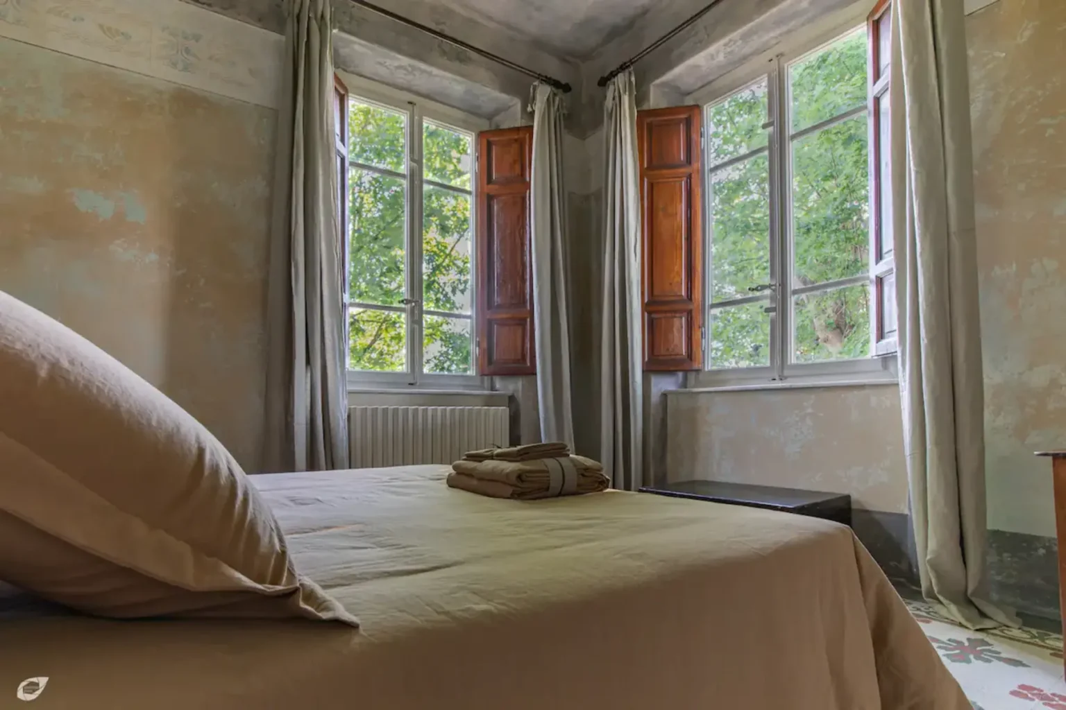 Cozy bedroom with a neutral-colored bedspread, a neatly folded towel, and large windows featuring wooden shutters that offer a view of greenery outside. Natural light floods the room, enhancing its inviting atmosphere. | Only on SopranoVillas