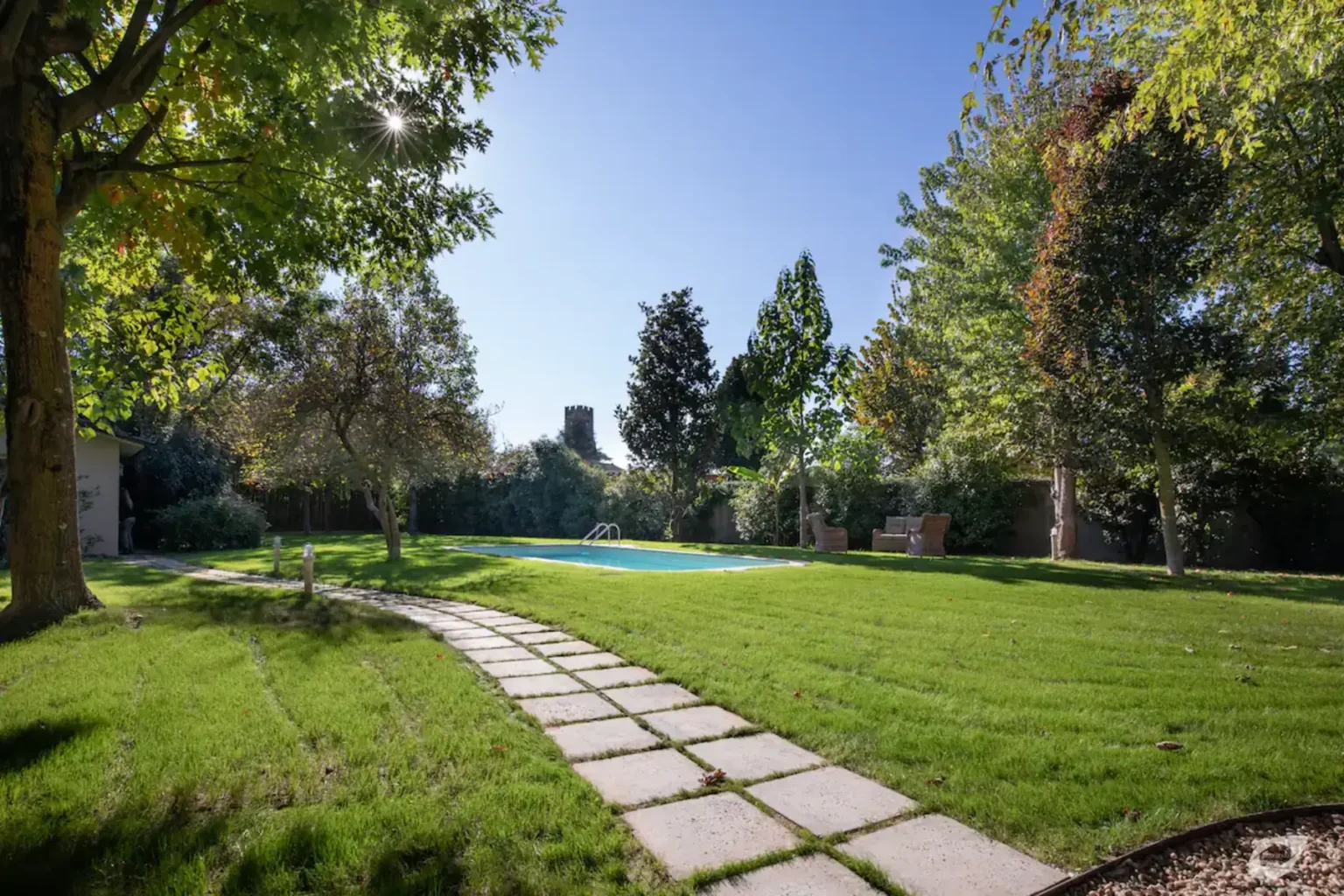 Lush green garden with a stone pathway leading to a swimming pool, surrounded by trees and clear blue sky. Ideal for outdoor relaxation and landscaping inspiration. | Only on SopranoVillas