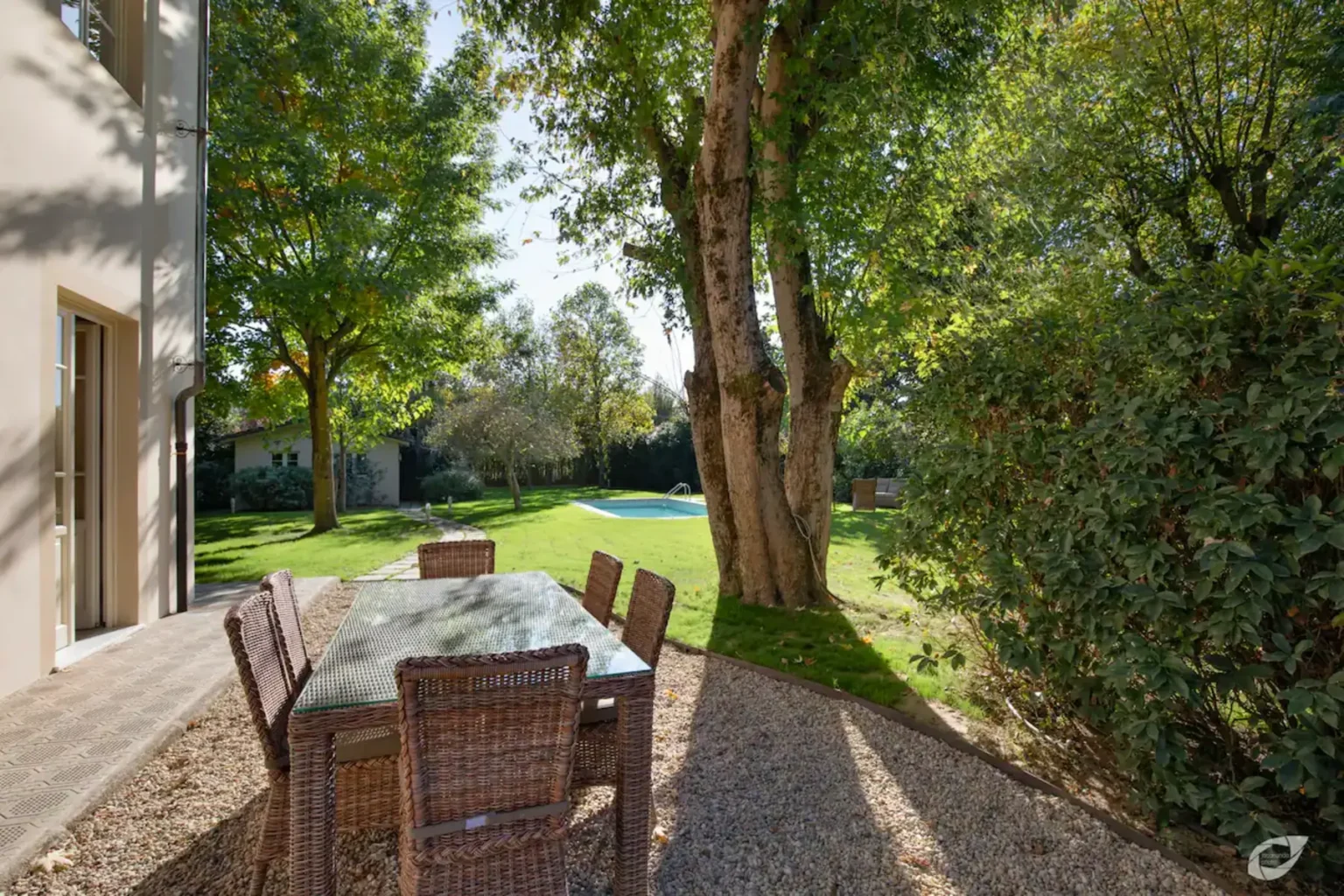 Outdoor dining area with a glass table and wicker chairs surrounded by lush greenery, featuring large trees and a view of a swimming pool in the background. Ideal for relaxing and entertaining in a serene garden setting. | Only on SopranoVillas