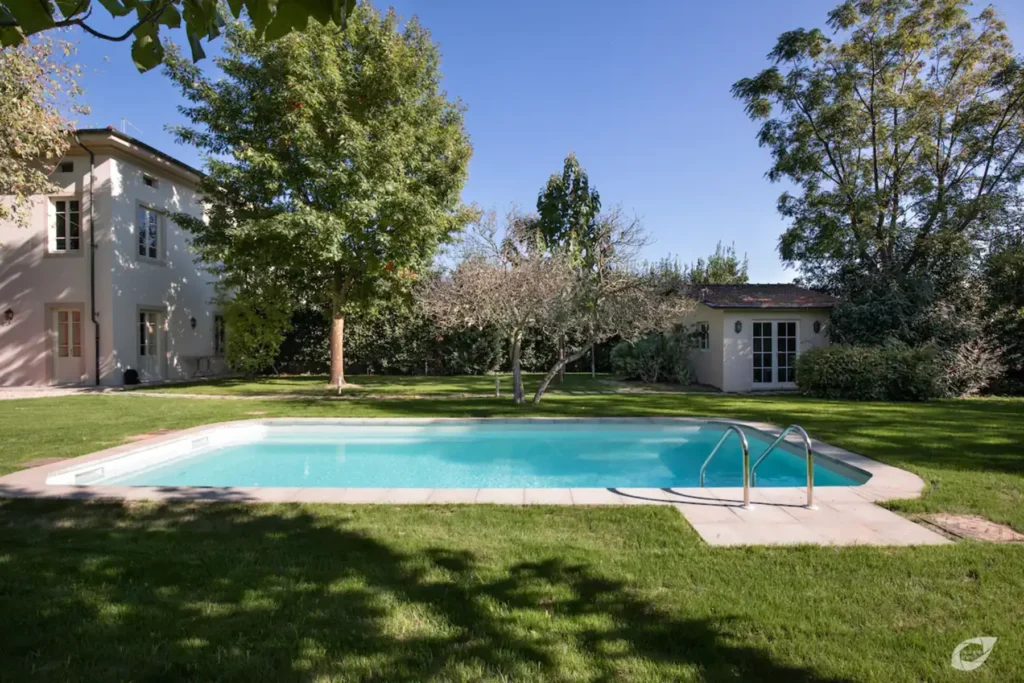 A serene backyard featuring a clear swimming pool surrounded by lush green grass and trees, with a modern home and a separate building in the background. Ideal for outdoor relaxation and entertaining. | Only on SopranoVillas