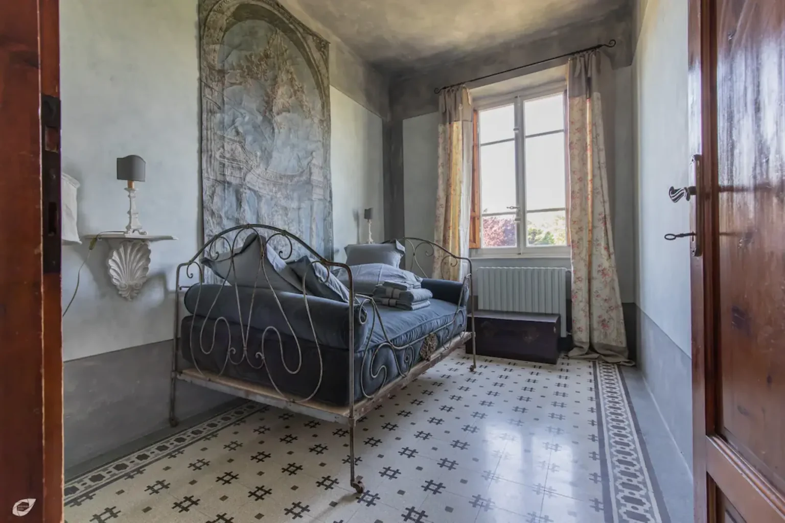 Cozy bedroom featuring an iron bed frame, patterned flooring, and large window with sheer curtains, showcasing a serene and inviting atmosphere. | Only on SopranoVillas