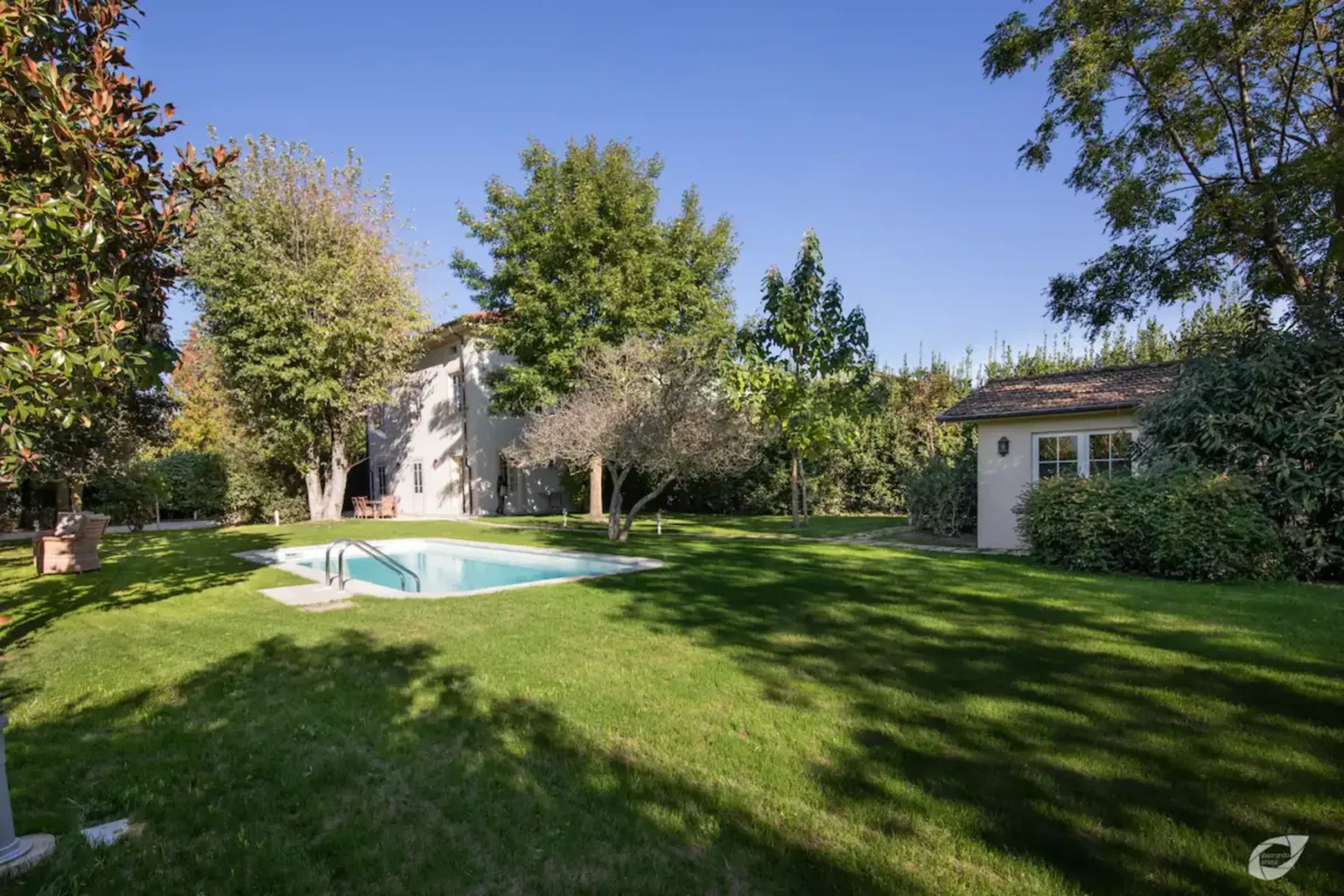 Spacious backyard featuring a swimming pool surrounded by lush green grass and trees, with a charming outbuilding visible in the background under a clear blue sky. Ideal for relaxation and outdoor activities. | Only on SopranoVillas