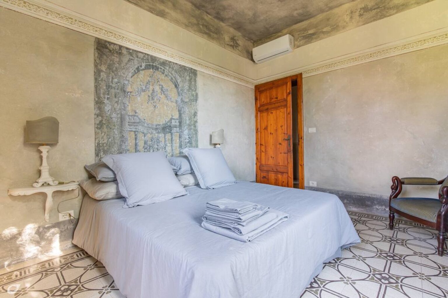 Cozy bedroom featuring a double bed with gray linens, decorative pillows, and a stack of towels. The room has textured walls, a wooden door, and a vintage chair, creating a warm and inviting atmosphere. | Only on SopranoVillas
