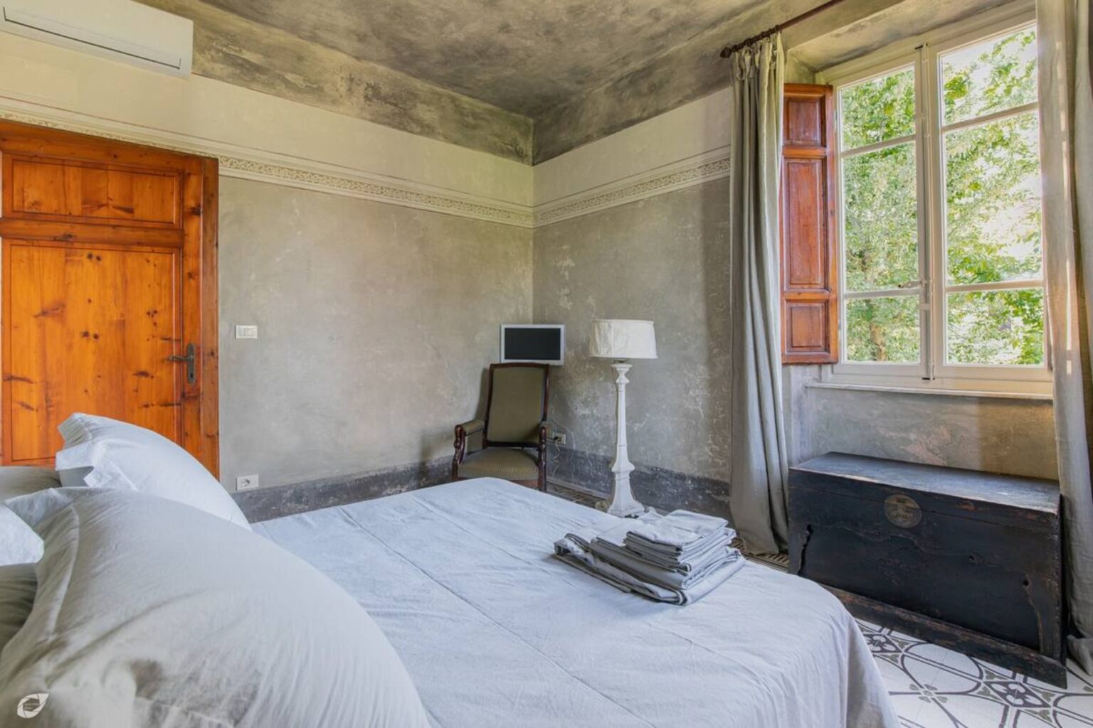 Cozy bedroom featuring a comfortable bed with neatly arranged linens, a stylish chair, a modern lamp, and a large window with natural light streaming in, showcasing a serene outdoor view. The room has a rustic wooden door and elegant decor elements, creating a warm and inviting atmosphere. | Only on SopranoVillas