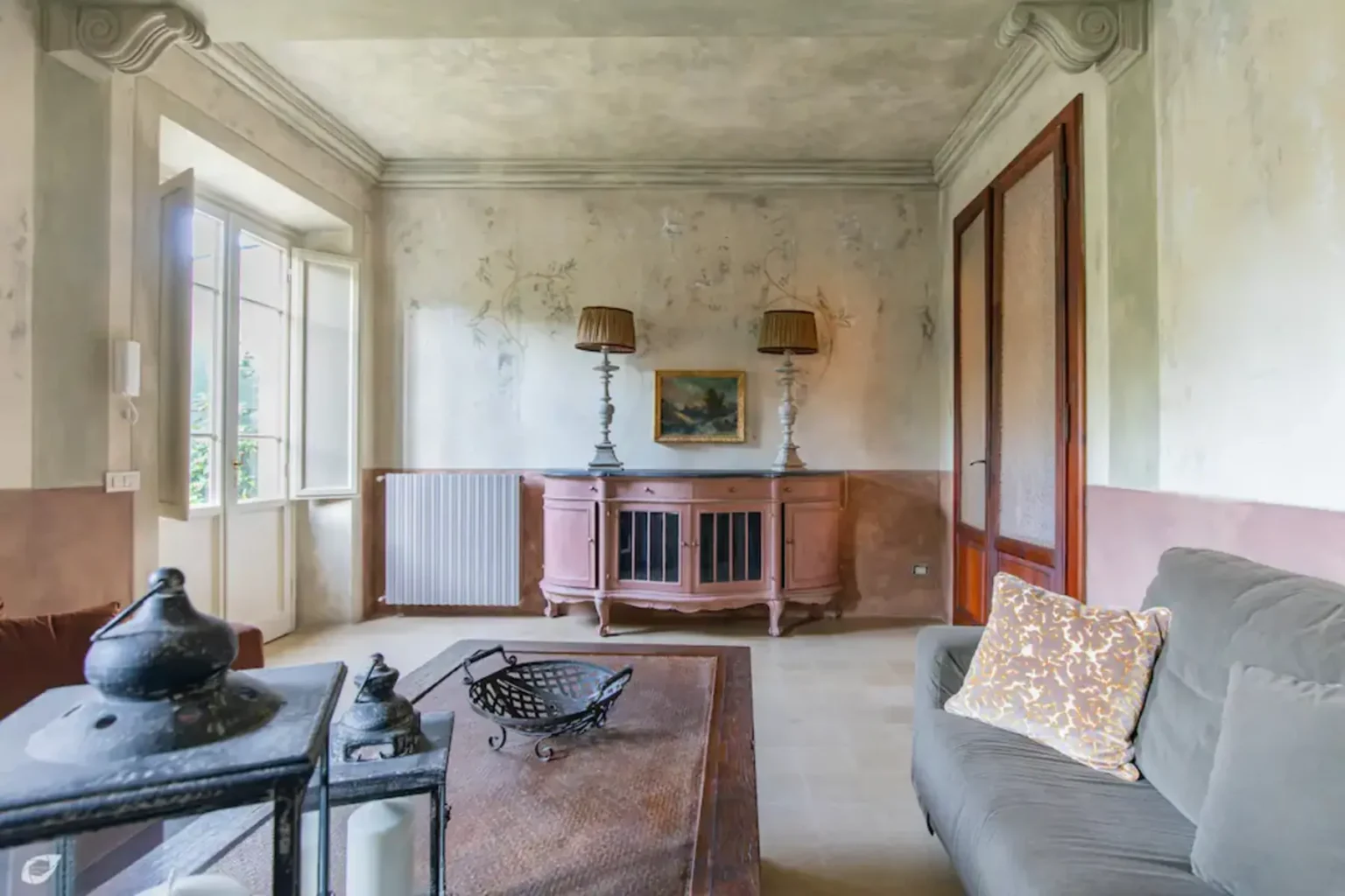 Interior of a vintage living room featuring a dusty, peeling wall, a wooden coffee table with decorative items, a gray sofa, and a classic radiator. Two lamps illuminate the space, and a small painting hangs above the radiator, creating a nostalgic atmosphere. | Only on SopranoVillas