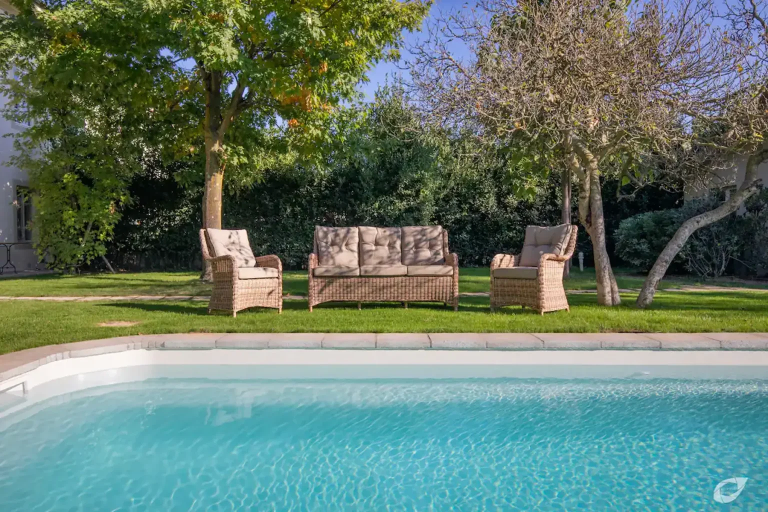 Cozy outdoor seating area with a sofa and two chairs beside a sparkling blue swimming pool, surrounded by lush greenery and trees. Ideal for relaxation and entertaining in a tranquil garden setting. | Only on SopranoVillas